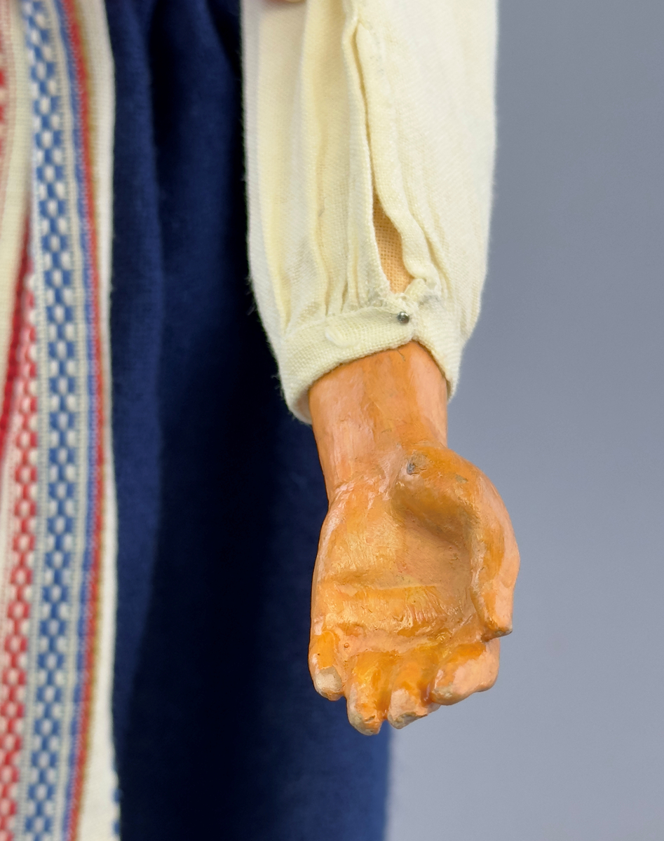 Docka klädd i kvinnlig sockendräkt från Ulleruds socken i Värmland. Kroppen är handsydd i linnetyg och stoppad med textilt material. Huvud och händer är av lergods. Kläder handsydda av handvävda tyger förutom skjorta, underkjol och halskläde, som är maskinvävt. Skjorta, broderad med myrgång runt sprund, har halsspjäll, halslinning och handlinning. Halsklädet, med tryck i gult, rosa och beige, hålls ihop av en sölja i nysilver med graverat mönster. Kjol sydd med lagda veck runtom, utom under förkläde, vars linning består av ett bomullsband. Förkläde, vävt i tuskaft och munkabälte, har lagda veck vid midja och bomullsband i rips till knytband. Livstycket är vändsytt/fodrat med ett rutigt bomullstyg och knäpps med 6 hyskar & hakar. Ullstrumporna är handstickade i slätstickning med en kant upptill i 2-2 resår. Huvudet är prytt med en vit linnehätta kantad med en knypplad linnespets. Dockans fötter bär svarta skinnskor.