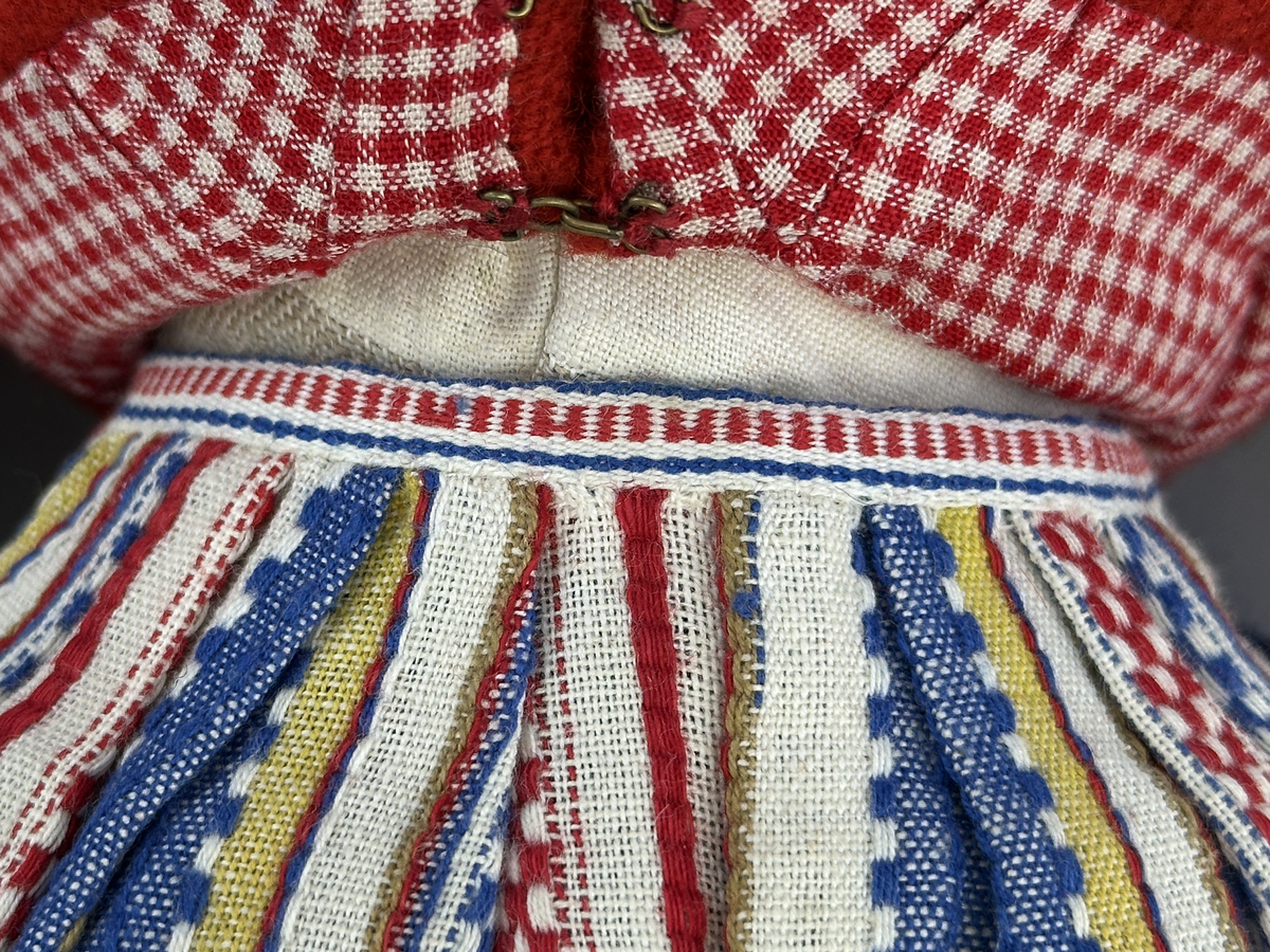 Docka klädd i kvinnlig sockendräkt från Ulleruds socken i Värmland. Kroppen är handsydd i linnetyg och stoppad med textilt material. Huvud och händer är av lergods. Kläder handsydda av handvävda tyger förutom skjorta, underkjol och halskläde, som är maskinvävt. Skjorta, broderad med myrgång runt sprund, har halsspjäll, halslinning och handlinning. Halsklädet, med tryck i gult, rosa och beige, hålls ihop av en sölja i nysilver med graverat mönster. Kjol sydd med lagda veck runtom, utom under förkläde, vars linning består av ett bomullsband. Förkläde, vävt i tuskaft och munkabälte, har lagda veck vid midja och bomullsband i rips till knytband. Livstycket är vändsytt/fodrat med ett rutigt bomullstyg och knäpps med 6 hyskar & hakar. Ullstrumporna är handstickade i slätstickning med en kant upptill i 2-2 resår. Huvudet är prytt med en vit linnehätta kantad med en knypplad linnespets. Dockans fötter bär svarta skinnskor.