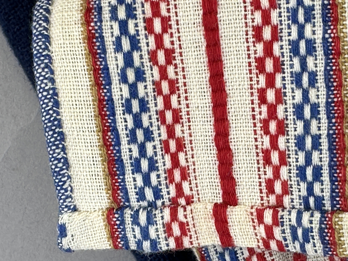 Docka klädd i kvinnlig sockendräkt från Ulleruds socken i Värmland. Kroppen är handsydd i linnetyg och stoppad med textilt material. Huvud och händer är av lergods. Kläder handsydda av handvävda tyger förutom skjorta, underkjol och halskläde, som är maskinvävt. Skjorta, broderad med myrgång runt sprund, har halsspjäll, halslinning och handlinning. Halsklädet, med tryck i gult, rosa och beige, hålls ihop av en sölja i nysilver med graverat mönster. Kjol sydd med lagda veck runtom, utom under förkläde, vars linning består av ett bomullsband. Förkläde, vävt i tuskaft och munkabälte, har lagda veck vid midja och bomullsband i rips till knytband. Livstycket är vändsytt/fodrat med ett rutigt bomullstyg och knäpps med 6 hyskar & hakar. Ullstrumporna är handstickade i slätstickning med en kant upptill i 2-2 resår. Huvudet är prytt med en vit linnehätta kantad med en knypplad linnespets. Dockans fötter bär svarta skinnskor.