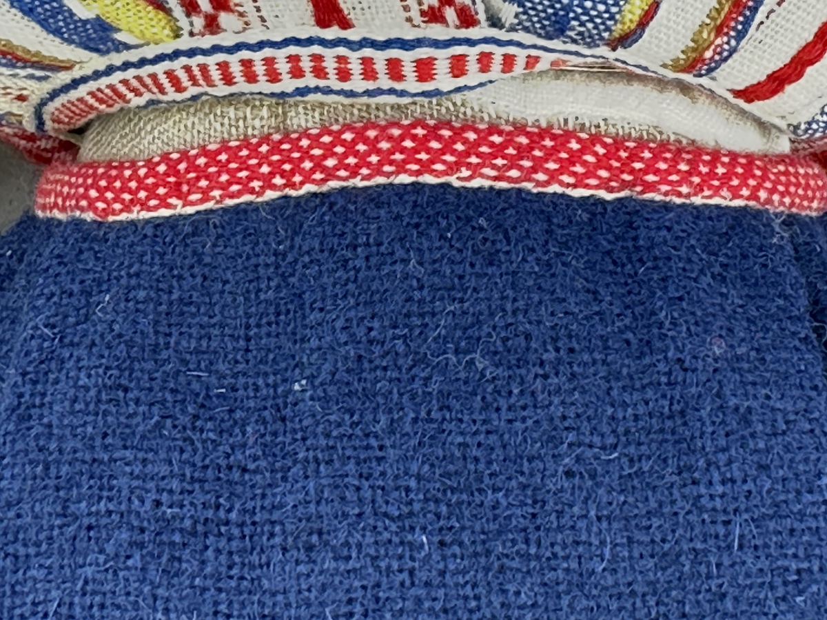 Docka klädd i kvinnlig sockendräkt från Ulleruds socken i Värmland. Kroppen är handsydd i linnetyg och stoppad med textilt material. Huvud och händer är av lergods. Kläder handsydda av handvävda tyger förutom skjorta, underkjol och halskläde, som är maskinvävt. Skjorta, broderad med myrgång runt sprund, har halsspjäll, halslinning och handlinning. Halsklädet, med tryck i gult, rosa och beige, hålls ihop av en sölja i nysilver med graverat mönster. Kjol sydd med lagda veck runtom, utom under förkläde, vars linning består av ett bomullsband. Förkläde, vävt i tuskaft och munkabälte, har lagda veck vid midja och bomullsband i rips till knytband. Livstycket är vändsytt/fodrat med ett rutigt bomullstyg och knäpps med 6 hyskar & hakar. Ullstrumporna är handstickade i slätstickning med en kant upptill i 2-2 resår. Huvudet är prytt med en vit linnehätta kantad med en knypplad linnespets. Dockans fötter bär svarta skinnskor.