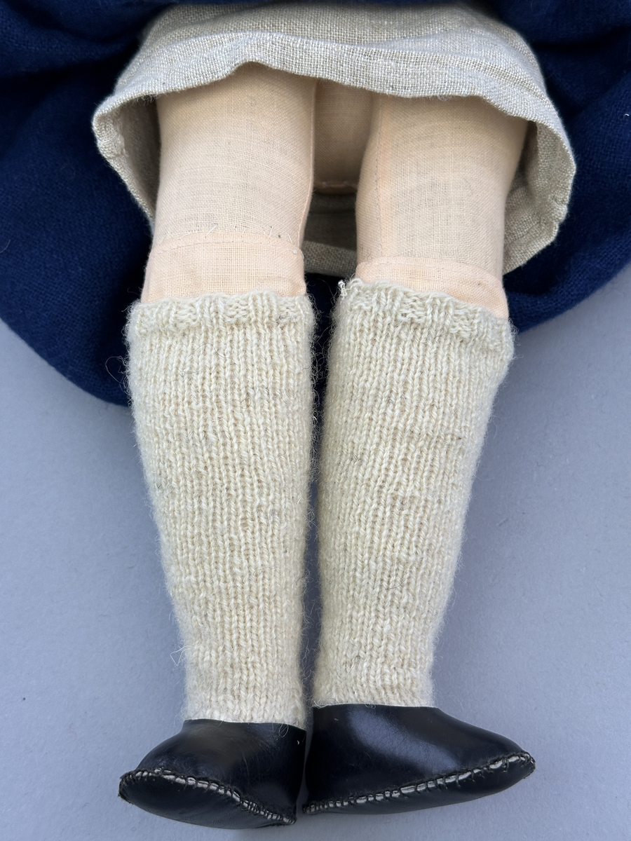 Docka klädd i kvinnlig sockendräkt från Ulleruds socken i Värmland. Kroppen är handsydd i linnetyg och stoppad med textilt material. Huvud och händer är av lergods. Kläder handsydda av handvävda tyger förutom skjorta, underkjol och halskläde, som är maskinvävt. Skjorta, broderad med myrgång runt sprund, har halsspjäll, halslinning och handlinning. Halsklädet, med tryck i gult, rosa och beige, hålls ihop av en sölja i nysilver med graverat mönster. Kjol sydd med lagda veck runtom, utom under förkläde, vars linning består av ett bomullsband. Förkläde, vävt i tuskaft och munkabälte, har lagda veck vid midja och bomullsband i rips till knytband. Livstycket är vändsytt/fodrat med ett rutigt bomullstyg och knäpps med 6 hyskar & hakar. Ullstrumporna är handstickade i slätstickning med en kant upptill i 2-2 resår. Huvudet är prytt med en vit linnehätta kantad med en knypplad linnespets. Dockans fötter bär svarta skinnskor.
