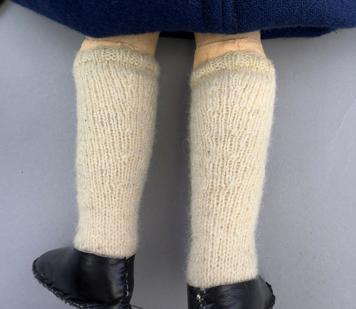 Docka klädd i kvinnlig sockendräkt från Ulleruds socken i Värmland. Kroppen är handsydd i linnetyg och stoppad med textilt material. Huvud och händer är av lergods. Kläder handsydda av handvävda tyger förutom skjorta, underkjol och halskläde, som är maskinvävt. Skjorta, broderad med myrgång runt sprund, har halsspjäll, halslinning och handlinning. Halsklädet, med tryck i gult, rosa och beige, hålls ihop av en sölja i nysilver med graverat mönster. Kjol sydd med lagda veck runtom, utom under förkläde, vars linning består av ett bomullsband. Förkläde, vävt i tuskaft och munkabälte, har lagda veck vid midja och bomullsband i rips till knytband. Livstycket är vändsytt/fodrat med ett rutigt bomullstyg och knäpps med 6 hyskar & hakar. Ullstrumporna är handstickade i slätstickning med en kant upptill i 2-2 resår. Huvudet är prytt med en vit linnehätta kantad med en knypplad linnespets. Dockans fötter bär svarta skinnskor.