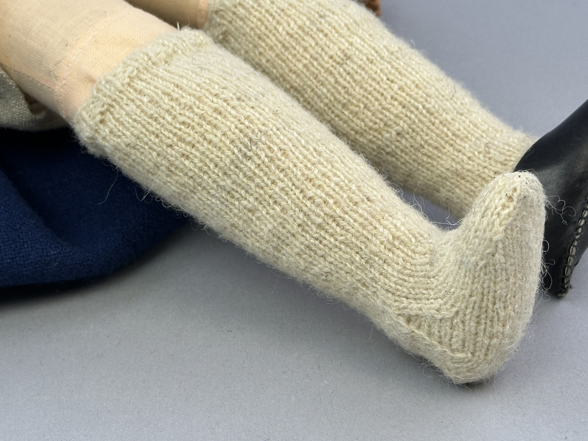 Docka klädd i kvinnlig sockendräkt från Ulleruds socken i Värmland. Kroppen är handsydd i linnetyg och stoppad med textilt material. Huvud och händer är av lergods. Kläder handsydda av handvävda tyger förutom skjorta, underkjol och halskläde, som är maskinvävt. Skjorta, broderad med myrgång runt sprund, har halsspjäll, halslinning och handlinning. Halsklädet, med tryck i gult, rosa och beige, hålls ihop av en sölja i nysilver med graverat mönster. Kjol sydd med lagda veck runtom, utom under förkläde, vars linning består av ett bomullsband. Förkläde, vävt i tuskaft och munkabälte, har lagda veck vid midja och bomullsband i rips till knytband. Livstycket är vändsytt/fodrat med ett rutigt bomullstyg och knäpps med 6 hyskar & hakar. Ullstrumporna är handstickade i slätstickning med en kant upptill i 2-2 resår. Huvudet är prytt med en vit linnehätta kantad med en knypplad linnespets. Dockans fötter bär svarta skinnskor.