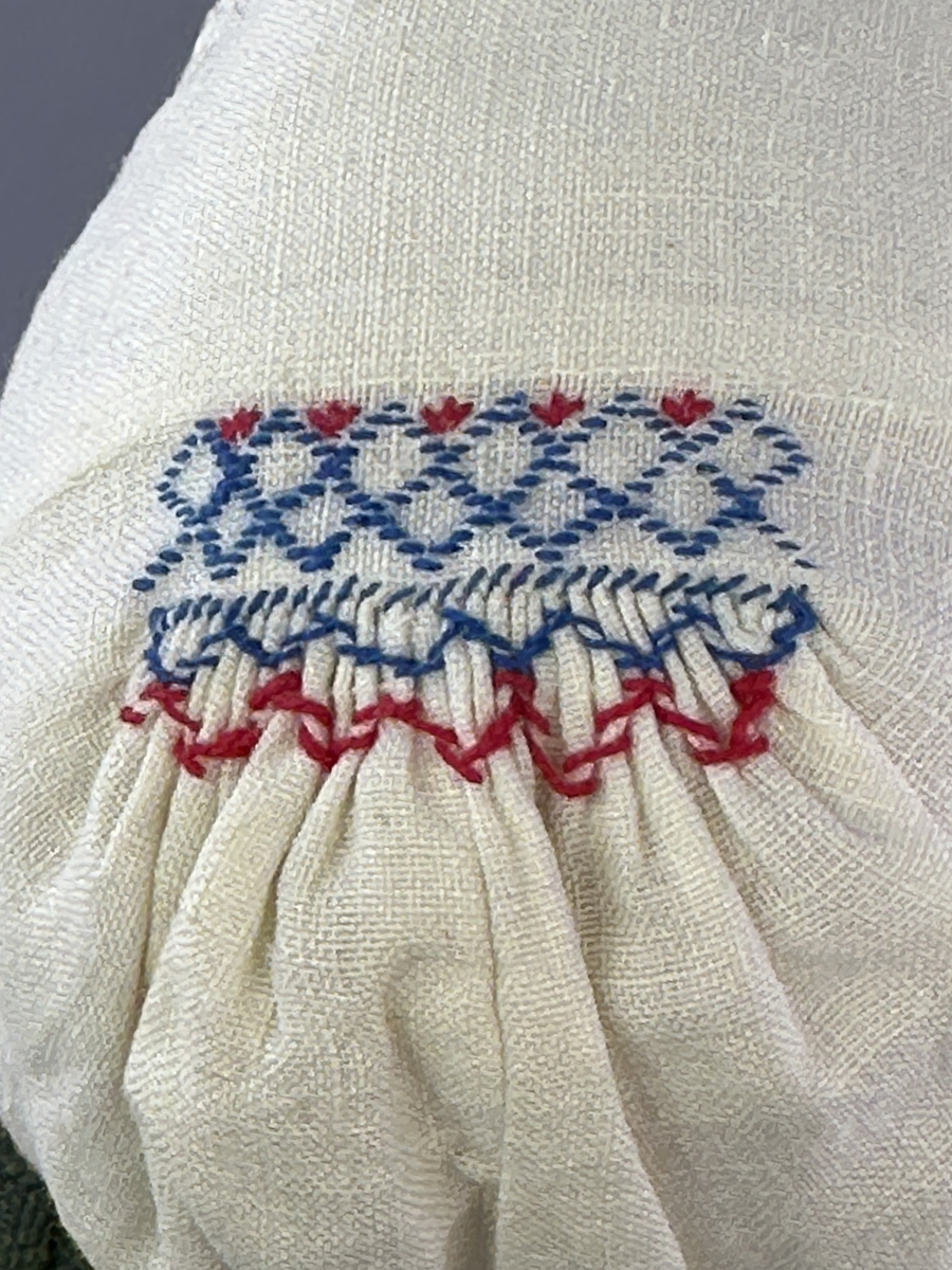 Docka klädd i kvinnlig sockendräkt från Dalby socken i Värmland. Kroppen är handsydd i linnetyg och stoppad med textilt material. Huvud och händer är av lergods. Kläder handsydda av handvävda tyger förutom skjorta, underkjol och halskläde, som är maskinvävt. Skjortan består av halsspjäll, krage och handlinning med stripade veck runt  krage och manschett. Skjortan är broderad med rutsöm vid sprund, krage, axelsöm och handlinning. Knytband bestående av trefläta (i rött och vitt) och tofsar (i rött, vitt och blått) pryder krage och manschett. Livkjol sydd med lagda veck runtom, utom under förkläde, har hals- och ärmhål kantad med passpoaler. Tyg till livkjol är vävt med bomullsvarp och ullinslag. Livet på livkjolen är vändsytt/fodrat med ett linnetyg och knäpps med 3 hyskar & hakar. Livkjolen är också dekorerad med en tre-fläta av rött ullgarn i nederkant. Även förklädet har lagda veck vid midja fastsydda mot en linning av bomullsband. Ullstrumporna är handstickade i slätstickning med en kant upptill i 2-2 resår samt en rad med avigmaskor, som "söm"-dekor på baksida ben. Huvudet är prytt med en vit linnehätta kantad med en knypplad linnespets samt ett broderi i blått och rött. Dockans fötter bär svarta skinnskor.

