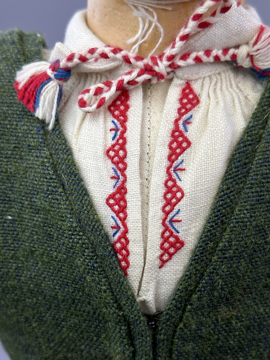 Docka klädd i kvinnlig sockendräkt från Dalby socken i Värmland. Kroppen är handsydd i linnetyg och stoppad med textilt material. Huvud och händer är av lergods. Kläder handsydda av handvävda tyger förutom skjorta, underkjol och halskläde, som är maskinvävt. Skjortan består av halsspjäll, krage och handlinning med stripade veck runt  krage och manschett. Skjortan är broderad med rutsöm vid sprund, krage, axelsöm och handlinning. Knytband bestående av trefläta (i rött och vitt) och tofsar (i rött, vitt och blått) pryder krage och manschett. Livkjol sydd med lagda veck runtom, utom under förkläde, har hals- och ärmhål kantad med passpoaler. Tyg till livkjol är vävt med bomullsvarp och ullinslag. Livet på livkjolen är vändsytt/fodrat med ett linnetyg och knäpps med 3 hyskar & hakar. Livkjolen är också dekorerad med en tre-fläta av rött ullgarn i nederkant. Även förklädet har lagda veck vid midja fastsydda mot en linning av bomullsband. Ullstrumporna är handstickade i slätstickning med en kant upptill i 2-2 resår samt en rad med avigmaskor, som "söm"-dekor på baksida ben. Huvudet är prytt med en vit linnehätta kantad med en knypplad linnespets samt ett broderi i blått och rött. Dockans fötter bär svarta skinnskor.

