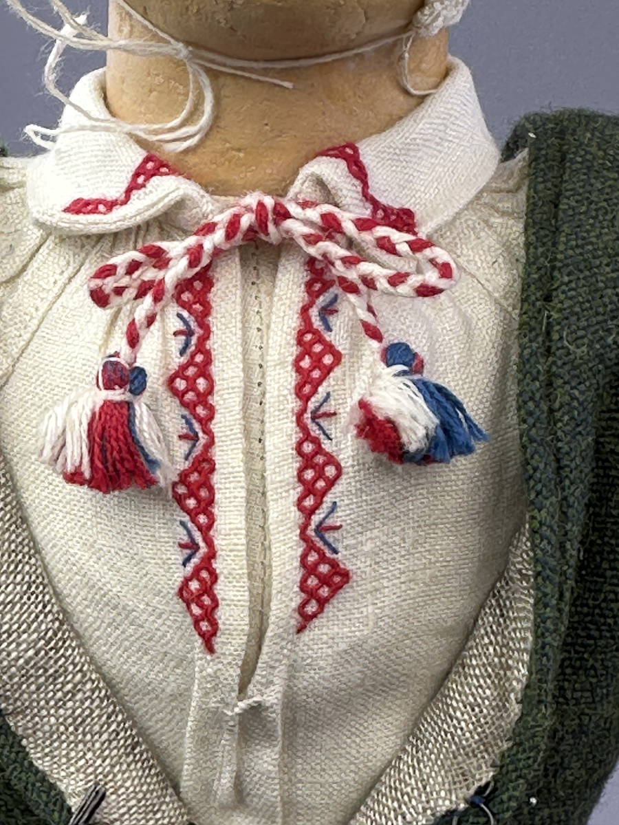 Docka klädd i kvinnlig sockendräkt från Dalby socken i Värmland. Kroppen är handsydd i linnetyg och stoppad med textilt material. Huvud och händer är av lergods. Kläder handsydda av handvävda tyger förutom skjorta, underkjol och halskläde, som är maskinvävt. Skjortan består av halsspjäll, krage och handlinning med stripade veck runt  krage och manschett. Skjortan är broderad med rutsöm vid sprund, krage, axelsöm och handlinning. Knytband bestående av trefläta (i rött och vitt) och tofsar (i rött, vitt och blått) pryder krage och manschett. Livkjol sydd med lagda veck runtom, utom under förkläde, har hals- och ärmhål kantad med passpoaler. Tyg till livkjol är vävt med bomullsvarp och ullinslag. Livet på livkjolen är vändsytt/fodrat med ett linnetyg och knäpps med 3 hyskar & hakar. Livkjolen är också dekorerad med en tre-fläta av rött ullgarn i nederkant. Även förklädet har lagda veck vid midja fastsydda mot en linning av bomullsband. Ullstrumporna är handstickade i slätstickning med en kant upptill i 2-2 resår samt en rad med avigmaskor, som "söm"-dekor på baksida ben. Huvudet är prytt med en vit linnehätta kantad med en knypplad linnespets samt ett broderi i blått och rött. Dockans fötter bär svarta skinnskor.

