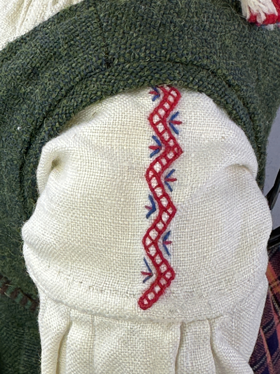 Docka klädd i kvinnlig sockendräkt från Dalby socken i Värmland. Kroppen är handsydd i linnetyg och stoppad med textilt material. Huvud och händer är av lergods. Kläder handsydda av handvävda tyger förutom skjorta, underkjol och halskläde, som är maskinvävt. Skjortan består av halsspjäll, krage och handlinning med stripade veck runt  krage och manschett. Skjortan är broderad med rutsöm vid sprund, krage, axelsöm och handlinning. Knytband bestående av trefläta (i rött och vitt) och tofsar (i rött, vitt och blått) pryder krage och manschett. Livkjol sydd med lagda veck runtom, utom under förkläde, har hals- och ärmhål kantad med passpoaler. Tyg till livkjol är vävt med bomullsvarp och ullinslag. Livet på livkjolen är vändsytt/fodrat med ett linnetyg och knäpps med 3 hyskar & hakar. Livkjolen är också dekorerad med en tre-fläta av rött ullgarn i nederkant. Även förklädet har lagda veck vid midja fastsydda mot en linning av bomullsband. Ullstrumporna är handstickade i slätstickning med en kant upptill i 2-2 resår samt en rad med avigmaskor, som "söm"-dekor på baksida ben. Huvudet är prytt med en vit linnehätta kantad med en knypplad linnespets samt ett broderi i blått och rött. Dockans fötter bär svarta skinnskor.


