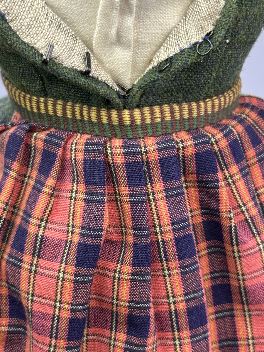 Docka klädd i kvinnlig sockendräkt från Dalby socken i Värmland. Kroppen är handsydd i linnetyg och stoppad med textilt material. Huvud och händer är av lergods. Kläder handsydda av handvävda tyger förutom skjorta, underkjol och halskläde, som är maskinvävt. Skjortan består av halsspjäll, krage och handlinning med stripade veck runt  krage och manschett. Skjortan är broderad med rutsöm vid sprund, krage, axelsöm och handlinning. Knytband bestående av trefläta (i rött och vitt) och tofsar (i rött, vitt och blått) pryder krage och manschett. Livkjol sydd med lagda veck runtom, utom under förkläde, har hals- och ärmhål kantad med passpoaler. Tyg till livkjol är vävt med bomullsvarp och ullinslag. Livet på livkjolen är vändsytt/fodrat med ett linnetyg och knäpps med 3 hyskar & hakar. Livkjolen är också dekorerad med en tre-fläta av rött ullgarn i nederkant. Även förklädet har lagda veck vid midja fastsydda mot en linning av bomullsband. Ullstrumporna är handstickade i slätstickning med en kant upptill i 2-2 resår samt en rad med avigmaskor, som "söm"-dekor på baksida ben. Huvudet är prytt med en vit linnehätta kantad med en knypplad linnespets samt ett broderi i blått och rött. Dockans fötter bär svarta skinnskor.

