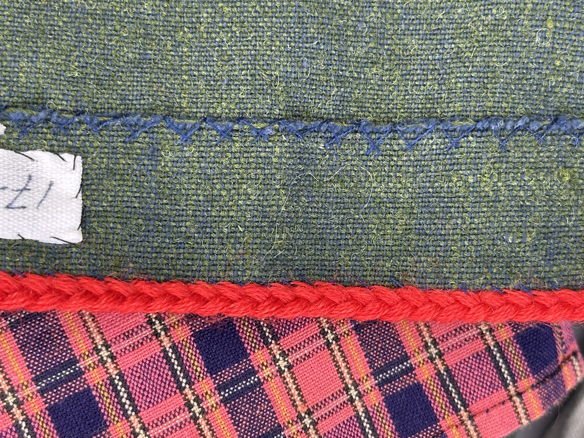 Docka klädd i kvinnlig sockendräkt från Dalby socken i Värmland. Kroppen är handsydd i linnetyg och stoppad med textilt material. Huvud och händer är av lergods. Kläder handsydda av handvävda tyger förutom skjorta, underkjol och halskläde, som är maskinvävt. Skjortan består av halsspjäll, krage och handlinning med stripade veck runt  krage och manschett. Skjortan är broderad med rutsöm vid sprund, krage, axelsöm och handlinning. Knytband bestående av trefläta (i rött och vitt) och tofsar (i rött, vitt och blått) pryder krage och manschett. Livkjol sydd med lagda veck runtom, utom under förkläde, har hals- och ärmhål kantad med passpoaler. Tyg till livkjol är vävt med bomullsvarp och ullinslag. Livet på livkjolen är vändsytt/fodrat med ett linnetyg och knäpps med 3 hyskar & hakar. Livkjolen är också dekorerad med en tre-fläta av rött ullgarn i nederkant. Även förklädet har lagda veck vid midja fastsydda mot en linning av bomullsband. Ullstrumporna är handstickade i slätstickning med en kant upptill i 2-2 resår samt en rad med avigmaskor, som "söm"-dekor på baksida ben. Huvudet är prytt med en vit linnehätta kantad med en knypplad linnespets samt ett broderi i blått och rött. Dockans fötter bär svarta skinnskor.

