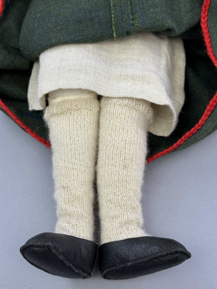 Docka klädd i kvinnlig sockendräkt från Dalby socken i Värmland. Kroppen är handsydd i linnetyg och stoppad med textilt material. Huvud och händer är av lergods. Kläder handsydda av handvävda tyger förutom skjorta, underkjol och halskläde, som är maskinvävt. Skjortan består av halsspjäll, krage och handlinning med stripade veck runt  krage och manschett. Skjortan är broderad med rutsöm vid sprund, krage, axelsöm och handlinning. Knytband bestående av trefläta (i rött och vitt) och tofsar (i rött, vitt och blått) pryder krage och manschett. Livkjol sydd med lagda veck runtom, utom under förkläde, har hals- och ärmhål kantad med passpoaler. Tyg till livkjol är vävt med bomullsvarp och ullinslag. Livet på livkjolen är vändsytt/fodrat med ett linnetyg och knäpps med 3 hyskar & hakar. Livkjolen är också dekorerad med en tre-fläta av rött ullgarn i nederkant. Även förklädet har lagda veck vid midja fastsydda mot en linning av bomullsband. Ullstrumporna är handstickade i slätstickning med en kant upptill i 2-2 resår samt en rad med avigmaskor, som "söm"-dekor på baksida ben. Huvudet är prytt med en vit linnehätta kantad med en knypplad linnespets samt ett broderi i blått och rött. Dockans fötter bär svarta skinnskor.

