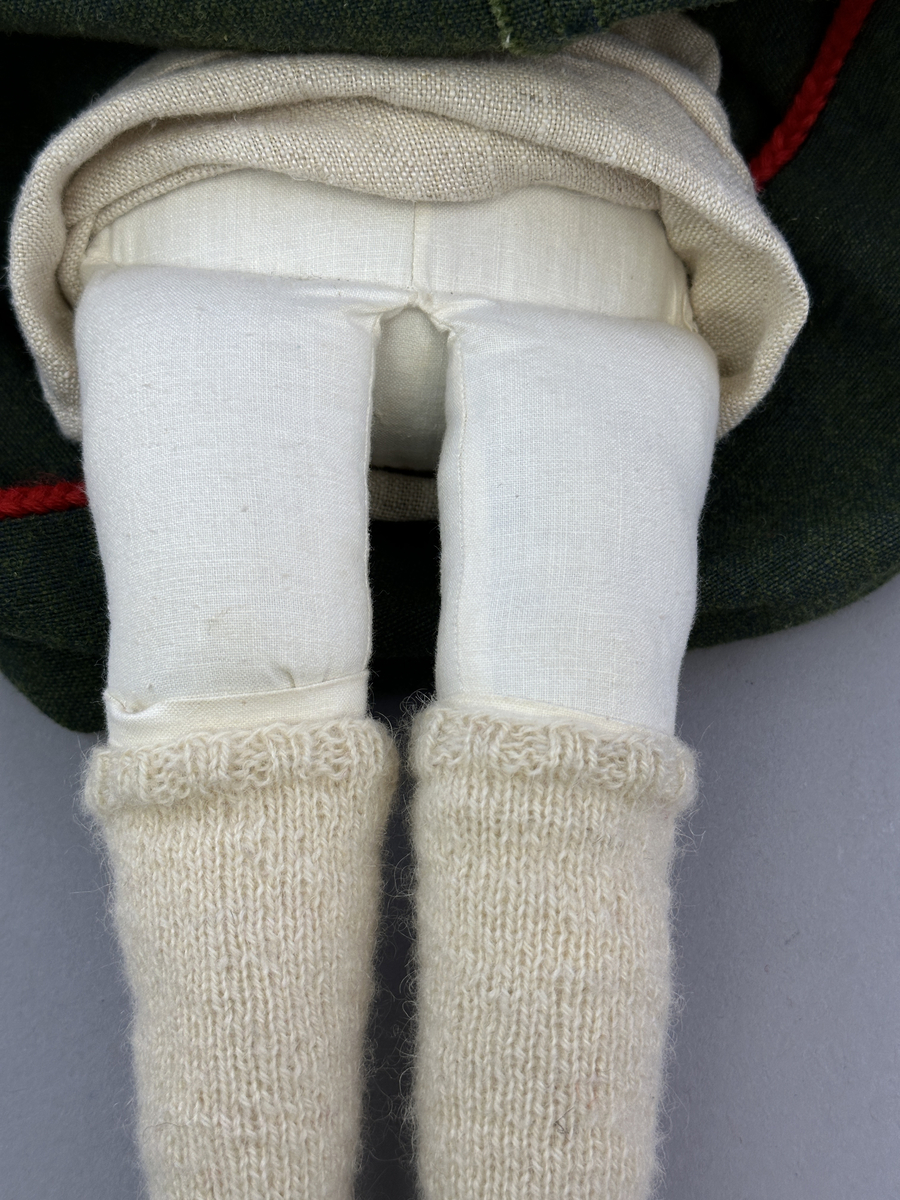 Docka klädd i kvinnlig sockendräkt från Dalby socken i Värmland. Kroppen är handsydd i linnetyg och stoppad med textilt material. Huvud och händer är av lergods. Kläder handsydda av handvävda tyger förutom skjorta, underkjol och halskläde, som är maskinvävt. Skjortan består av halsspjäll, krage och handlinning med stripade veck runt  krage och manschett. Skjortan är broderad med rutsöm vid sprund, krage, axelsöm och handlinning. Knytband bestående av trefläta (i rött och vitt) och tofsar (i rött, vitt och blått) pryder krage och manschett. Livkjol sydd med lagda veck runtom, utom under förkläde, har hals- och ärmhål kantad med passpoaler. Tyg till livkjol är vävt med bomullsvarp och ullinslag. Livet på livkjolen är vändsytt/fodrat med ett linnetyg och knäpps med 3 hyskar & hakar. Livkjolen är också dekorerad med en tre-fläta av rött ullgarn i nederkant. Även förklädet har lagda veck vid midja fastsydda mot en linning av bomullsband. Ullstrumporna är handstickade i slätstickning med en kant upptill i 2-2 resår samt en rad med avigmaskor, som "söm"-dekor på baksida ben. Huvudet är prytt med en vit linnehätta kantad med en knypplad linnespets samt ett broderi i blått och rött. Dockans fötter bär svarta skinnskor.

