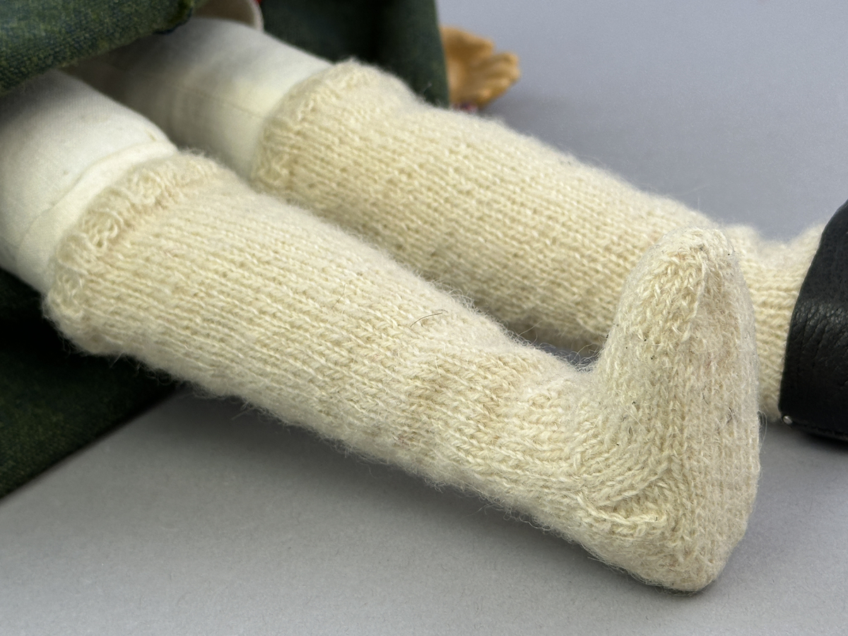 Docka klädd i kvinnlig sockendräkt från Dalby socken i Värmland. Kroppen är handsydd i linnetyg och stoppad med textilt material. Huvud och händer är av lergods. Kläder handsydda av handvävda tyger förutom skjorta, underkjol och halskläde, som är maskinvävt. Skjortan består av halsspjäll, krage och handlinning med stripade veck runt  krage och manschett. Skjortan är broderad med rutsöm vid sprund, krage, axelsöm och handlinning. Knytband bestående av trefläta (i rött och vitt) och tofsar (i rött, vitt och blått) pryder krage och manschett. Livkjol sydd med lagda veck runtom, utom under förkläde, har hals- och ärmhål kantad med passpoaler. Tyg till livkjol är vävt med bomullsvarp och ullinslag. Livet på livkjolen är vändsytt/fodrat med ett linnetyg och knäpps med 3 hyskar & hakar. Livkjolen är också dekorerad med en tre-fläta av rött ullgarn i nederkant. Även förklädet har lagda veck vid midja fastsydda mot en linning av bomullsband. Ullstrumporna är handstickade i slätstickning med en kant upptill i 2-2 resår samt en rad med avigmaskor, som "söm"-dekor på baksida ben. Huvudet är prytt med en vit linnehätta kantad med en knypplad linnespets samt ett broderi i blått och rött. Dockans fötter bär svarta skinnskor.

