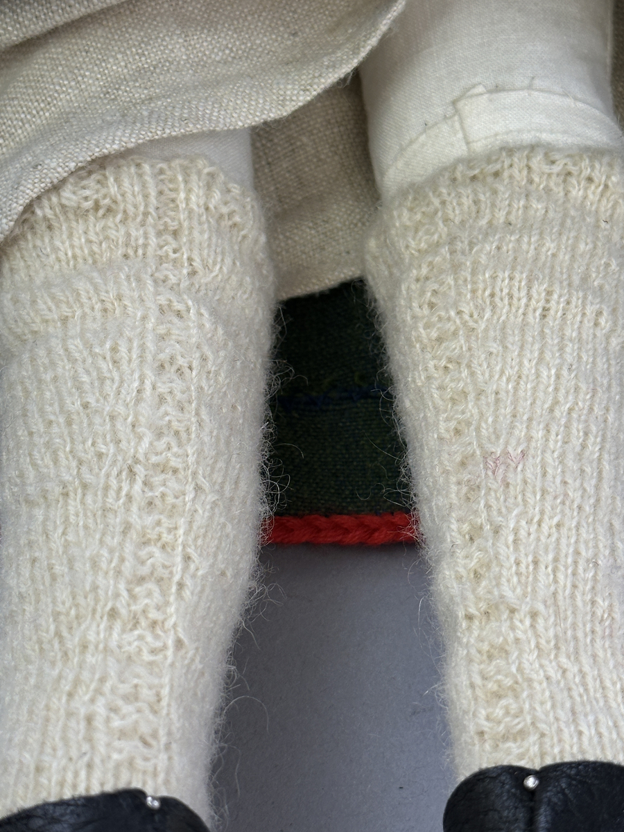 Docka klädd i kvinnlig sockendräkt från Dalby socken i Värmland. Kroppen är handsydd i linnetyg och stoppad med textilt material. Huvud och händer är av lergods. Kläder handsydda av handvävda tyger förutom skjorta, underkjol och halskläde, som är maskinvävt. Skjortan består av halsspjäll, krage och handlinning med stripade veck runt  krage och manschett. Skjortan är broderad med rutsöm vid sprund, krage, axelsöm och handlinning. Knytband bestående av trefläta (i rött och vitt) och tofsar (i rött, vitt och blått) pryder krage och manschett. Livkjol sydd med lagda veck runtom, utom under förkläde, har hals- och ärmhål kantad med passpoaler. Tyg till livkjol är vävt med bomullsvarp och ullinslag. Livet på livkjolen är vändsytt/fodrat med ett linnetyg och knäpps med 3 hyskar & hakar. Livkjolen är också dekorerad med en tre-fläta av rött ullgarn i nederkant. Även förklädet har lagda veck vid midja fastsydda mot en linning av bomullsband. Ullstrumporna är handstickade i slätstickning med en kant upptill i 2-2 resår samt en rad med avigmaskor, som "söm"-dekor på baksida ben. Huvudet är prytt med en vit linnehätta kantad med en knypplad linnespets samt ett broderi i blått och rött. Dockans fötter bär svarta skinnskor.

