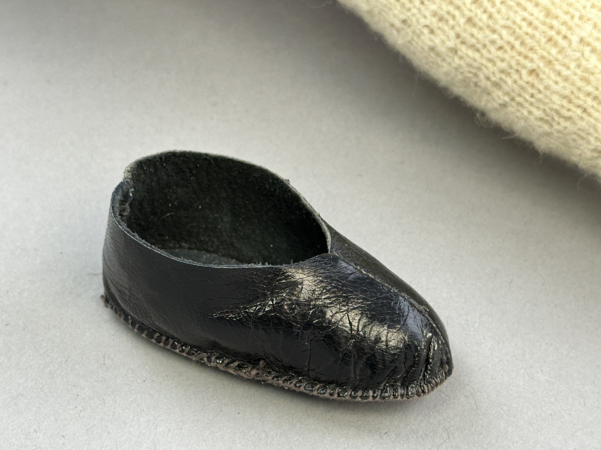 Docka klädd i kvinnlig sockendräkt från Dalby socken i Värmland. Kroppen är handsydd i linnetyg och stoppad med textilt material. Huvud och händer är av lergods. Kläder handsydda av handvävda tyger förutom skjorta, underkjol och halskläde, som är maskinvävt. Skjortan består av halsspjäll, krage och handlinning med stripade veck runt  krage och manschett. Skjortan är broderad med rutsöm vid sprund, krage, axelsöm och handlinning. Knytband bestående av trefläta (i rött och vitt) och tofsar (i rött, vitt och blått) pryder krage och manschett. Livkjol sydd med lagda veck runtom, utom under förkläde, har hals- och ärmhål kantad med passpoaler. Tyg till livkjol är vävt med bomullsvarp och ullinslag. Livet på livkjolen är vändsytt/fodrat med ett linnetyg och knäpps med 3 hyskar & hakar. Livkjolen är också dekorerad med en tre-fläta av rött ullgarn i nederkant. Även förklädet har lagda veck vid midja fastsydda mot en linning av bomullsband. Ullstrumporna är handstickade i slätstickning med en kant upptill i 2-2 resår samt en rad med avigmaskor, som "söm"-dekor på baksida ben. Huvudet är prytt med en vit linnehätta kantad med en knypplad linnespets samt ett broderi i blått och rött. Dockans fötter bär svarta skinnskor.

