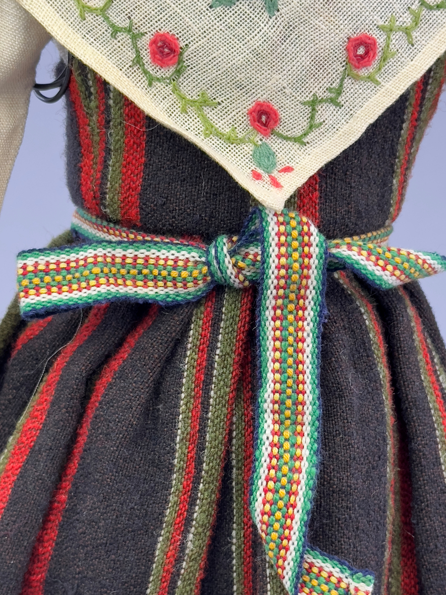 Docka klädd i kvinnlig sockendräkt från Fryksdals härad i Värmland. Kroppen är handsydd i linnetyg och stoppad med textilt material. Huvud och händer är av lergods. Kläder handsydda av handvävda tyger förutom skjorta, underkjol och halskläde, som är maskinvävt. Halskläde är broderat med tambursöm i gult, rosa, vinrött och grönt i 3 olika nyanser. Skjortan har halsspjäll, halslinning och stripade veck runt halskrage. Livkjol sydd med lagda veck runtom, utom under förkläde. Tyget till livkjol och förkläde är vävt med bomullsvarp och ullinslag. Även förklädet har lagda veck vid midja fastsydda mot en linning av bomullsband. Livet på livkjolen är vändsytt/fodrat med ett linnetyg och knäpps med 5 hyskar & hakar. Ullstrumporna är handstickade i slätstickning med en kant upptill i 2-2 resår samt en rad med avigmaskor, som "söm"-dekor på baksida ben.
