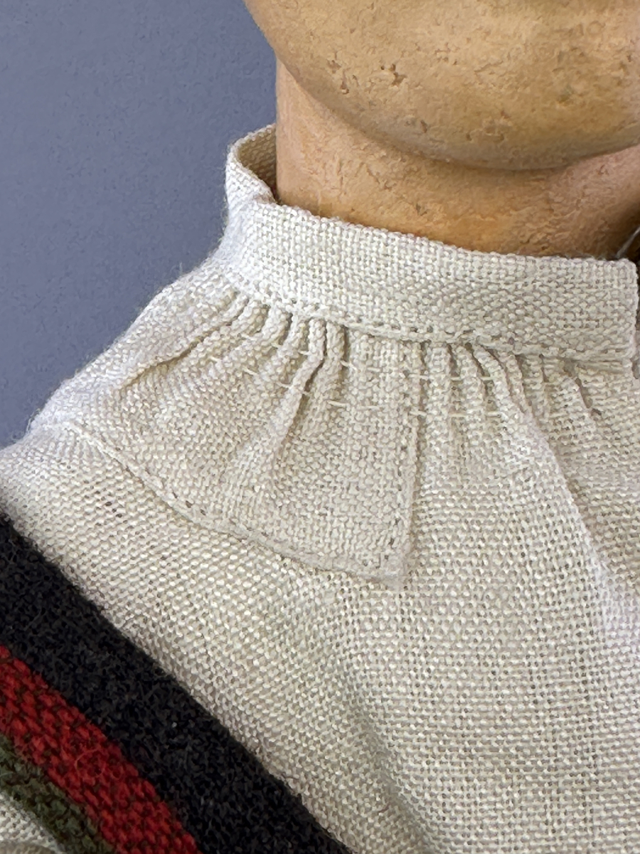 Docka klädd i kvinnlig sockendräkt från Fryksdals härad i Värmland. Kroppen är handsydd i linnetyg och stoppad med textilt material. Huvud och händer är av lergods. Kläder handsydda av handvävda tyger förutom skjorta, underkjol och halskläde, som är maskinvävt. Halskläde är broderat med tambursöm i gult, rosa, vinrött och grönt i 3 olika nyanser. Skjortan har halsspjäll, halslinning och stripade veck runt halskrage. Livkjol sydd med lagda veck runtom, utom under förkläde. Tyget till livkjol och förkläde är vävt med bomullsvarp och ullinslag. Även förklädet har lagda veck vid midja fastsydda mot en linning av bomullsband. Livet på livkjolen är vändsytt/fodrat med ett linnetyg och knäpps med 5 hyskar & hakar. Ullstrumporna är handstickade i slätstickning med en kant upptill i 2-2 resår samt en rad med avigmaskor, som "söm"-dekor på baksida ben.