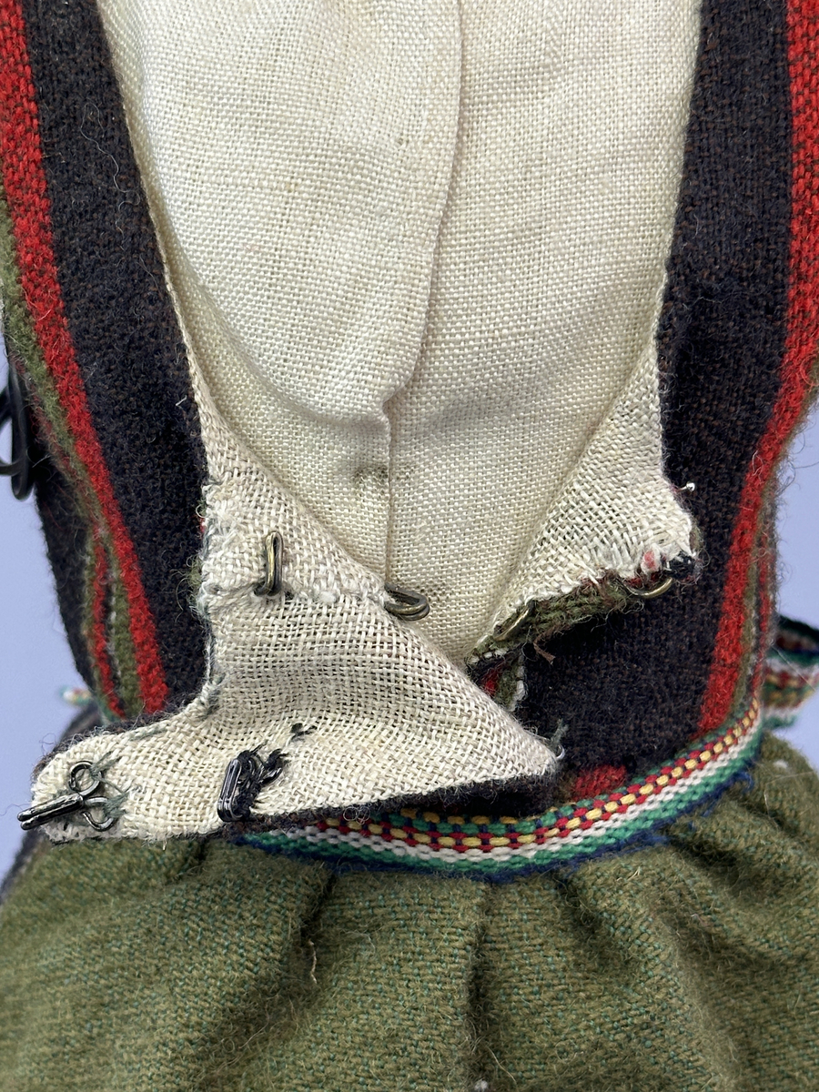 Docka klädd i kvinnlig sockendräkt från Fryksdals härad i Värmland. Kroppen är handsydd i linnetyg och stoppad med textilt material. Huvud och händer är av lergods. Kläder handsydda av handvävda tyger förutom skjorta, underkjol och halskläde, som är maskinvävt. Halskläde är broderat med tambursöm i gult, rosa, vinrött och grönt i 3 olika nyanser. Skjortan har halsspjäll, halslinning och stripade veck runt halskrage. Livkjol sydd med lagda veck runtom, utom under förkläde. Tyget till livkjol och förkläde är vävt med bomullsvarp och ullinslag. Även förklädet har lagda veck vid midja fastsydda mot en linning av bomullsband. Livet på livkjolen är vändsytt/fodrat med ett linnetyg och knäpps med 5 hyskar & hakar. Ullstrumporna är handstickade i slätstickning med en kant upptill i 2-2 resår samt en rad med avigmaskor, som "söm"-dekor på baksida ben.