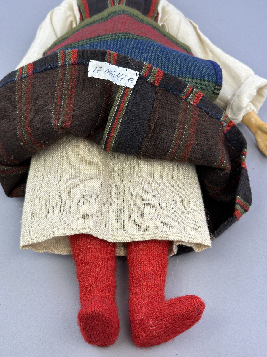 Docka klädd i kvinnlig sockendräkt från Fryksdals härad i Värmland. Kroppen är handsydd i linnetyg och stoppad med textilt material. Huvud och händer är av lergods. Kläder handsydda av handvävda tyger förutom skjorta, underkjol och halskläde, som är maskinvävt. Halskläde är broderat med tambursöm i gult, rosa, vinrött och grönt i 3 olika nyanser. Skjortan har halsspjäll, halslinning och stripade veck runt halskrage. Livkjol sydd med lagda veck runtom, utom under förkläde. Tyget till livkjol och förkläde är vävt med bomullsvarp och ullinslag. Även förklädet har lagda veck vid midja fastsydda mot en linning av bomullsband. Livet på livkjolen är vändsytt/fodrat med ett linnetyg och knäpps med 5 hyskar & hakar. Ullstrumporna är handstickade i slätstickning med en kant upptill i 2-2 resår samt en rad med avigmaskor, som "söm"-dekor på baksida ben.