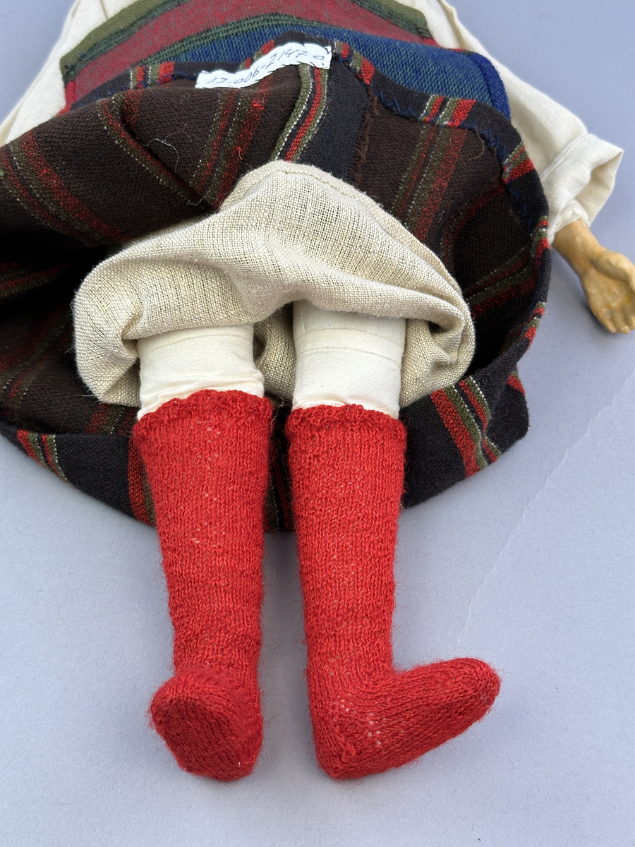 Docka klädd i kvinnlig sockendräkt från Fryksdals härad i Värmland. Kroppen är handsydd i linnetyg och stoppad med textilt material. Huvud och händer är av lergods. Kläder handsydda av handvävda tyger förutom skjorta, underkjol och halskläde, som är maskinvävt. Halskläde är broderat med tambursöm i gult, rosa, vinrött och grönt i 3 olika nyanser. Skjortan har halsspjäll, halslinning och stripade veck runt halskrage. Livkjol sydd med lagda veck runtom, utom under förkläde. Tyget till livkjol och förkläde är vävt med bomullsvarp och ullinslag. Även förklädet har lagda veck vid midja fastsydda mot en linning av bomullsband. Livet på livkjolen är vändsytt/fodrat med ett linnetyg och knäpps med 5 hyskar & hakar. Ullstrumporna är handstickade i slätstickning med en kant upptill i 2-2 resår samt en rad med avigmaskor, som "söm"-dekor på baksida ben.
