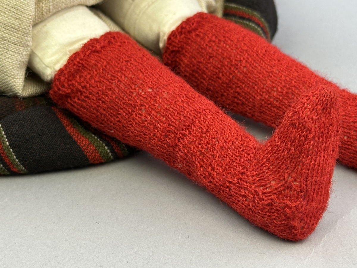 Docka klädd i kvinnlig sockendräkt från Fryksdals härad i Värmland. Kroppen är handsydd i linnetyg och stoppad med textilt material. Huvud och händer är av lergods. Kläder handsydda av handvävda tyger förutom skjorta, underkjol och halskläde, som är maskinvävt. Halskläde är broderat med tambursöm i gult, rosa, vinrött och grönt i 3 olika nyanser. Skjortan har halsspjäll, halslinning och stripade veck runt halskrage. Livkjol sydd med lagda veck runtom, utom under förkläde. Tyget till livkjol och förkläde är vävt med bomullsvarp och ullinslag. Även förklädet har lagda veck vid midja fastsydda mot en linning av bomullsband. Livet på livkjolen är vändsytt/fodrat med ett linnetyg och knäpps med 5 hyskar & hakar. Ullstrumporna är handstickade i slätstickning med en kant upptill i 2-2 resår samt en rad med avigmaskor, som "söm"-dekor på baksida ben.