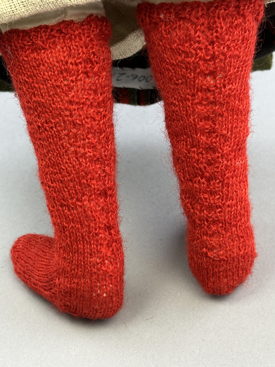 Docka klädd i kvinnlig sockendräkt från Fryksdals härad i Värmland. Kroppen är handsydd i linnetyg och stoppad med textilt material. Huvud och händer är av lergods. Kläder handsydda av handvävda tyger förutom skjorta, underkjol och halskläde, som är maskinvävt. Halskläde är broderat med tambursöm i gult, rosa, vinrött och grönt i 3 olika nyanser. Skjortan har halsspjäll, halslinning och stripade veck runt halskrage. Livkjol sydd med lagda veck runtom, utom under förkläde. Tyget till livkjol och förkläde är vävt med bomullsvarp och ullinslag. Även förklädet har lagda veck vid midja fastsydda mot en linning av bomullsband. Livet på livkjolen är vändsytt/fodrat med ett linnetyg och knäpps med 5 hyskar & hakar. Ullstrumporna är handstickade i slätstickning med en kant upptill i 2-2 resår samt en rad med avigmaskor, som "söm"-dekor på baksida ben.