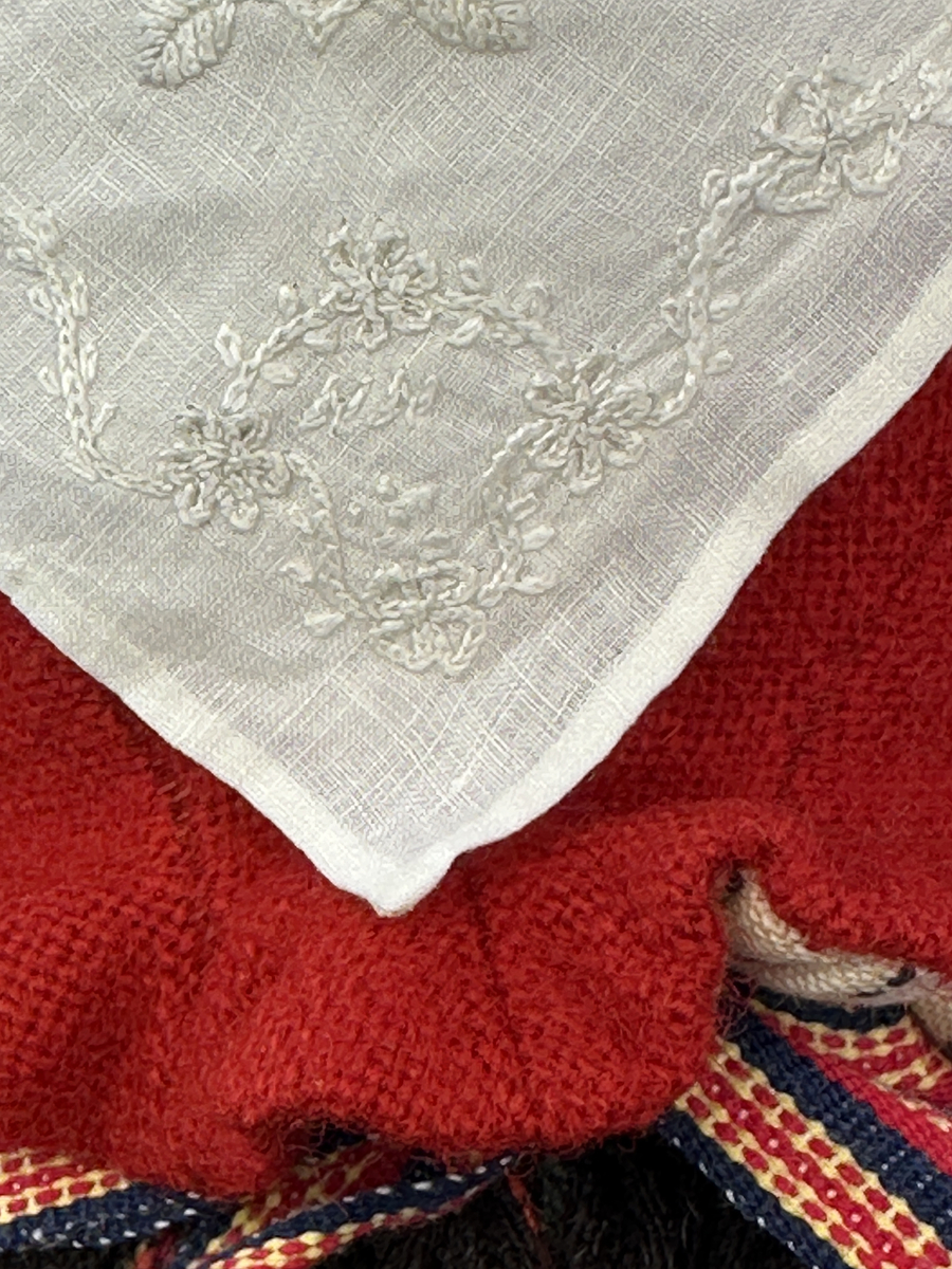 Docka klädd i kvinnlig sockendräkt från Sunne socken i Värmland. Kroppen är handsydd i linnetyg och stoppad med textilt material. Huvud och händer är av lergods. Kläder handsydda av handvävda tyger förutom skjorta, underkjol och halskläde, som är maskinvävt. Skjorta, broderad med stopphålsömsbroderi i vitt lingarn längs sprund mitt fram, har stripade veck runt krage och manschett. Halskläde, broderad med tambursöm i vitt moulinegarn, hålls ihop av en sölja i nysilver med graverat mönster. Yllekjol är rynkad upptill, utom under förkläde, vars linning och skoningsband består av ett bomullsband. Förklädet har lagda veck vid midja fastsydda mot en linning av bomullsband. Livstycket med sköld i ylletyg, är vändsytt/fodrat med ett mönstrat bomullstyg och knäpps med 5 hyskar & hakar samt har 2 små tennknappar fastsydda på utsidan. Ullstrumporna är handstickade i slätstickning med en kant upptill i 2-2 resår. Huvudet är prytt med en vit linnehätta kantad med en knypplad linnespets. Dockans fötter bär svarta skinnskor.