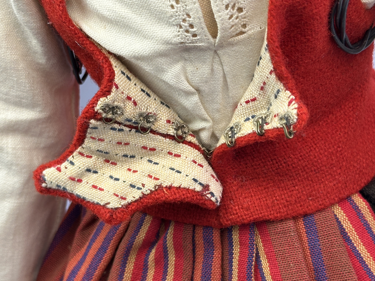 Docka klädd i kvinnlig sockendräkt från Sunne socken i Värmland. Kroppen är handsydd i linnetyg och stoppad med textilt material. Huvud och händer är av lergods. Kläder handsydda av handvävda tyger förutom skjorta, underkjol och halskläde, som är maskinvävt. Skjorta, broderad med stopphålsömsbroderi i vitt lingarn längs sprund mitt fram, har stripade veck runt krage och manschett. Halskläde, broderad med tambursöm i vitt moulinegarn, hålls ihop av en sölja i nysilver med graverat mönster. Yllekjol är rynkad upptill, utom under förkläde, vars linning och skoningsband består av ett bomullsband. Förklädet har lagda veck vid midja fastsydda mot en linning av bomullsband. Livstycket med sköld i ylletyg, är vändsytt/fodrat med ett mönstrat bomullstyg och knäpps med 5 hyskar & hakar samt har 2 små tennknappar fastsydda på utsidan. Ullstrumporna är handstickade i slätstickning med en kant upptill i 2-2 resår. Huvudet är prytt med en vit linnehätta kantad med en knypplad linnespets. Dockans fötter bär svarta skinnskor.