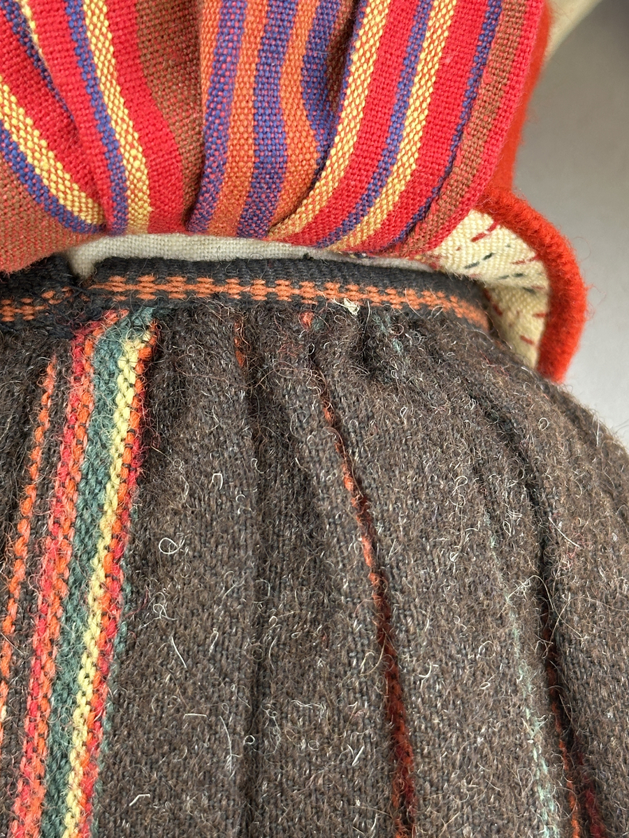 Docka klädd i kvinnlig sockendräkt från Sunne socken i Värmland. Kroppen är handsydd i linnetyg och stoppad med textilt material. Huvud och händer är av lergods. Kläder handsydda av handvävda tyger förutom skjorta, underkjol och halskläde, som är maskinvävt. Skjorta, broderad med stopphålsömsbroderi i vitt lingarn längs sprund mitt fram, har stripade veck runt krage och manschett. Halskläde, broderad med tambursöm i vitt moulinegarn, hålls ihop av en sölja i nysilver med graverat mönster. Yllekjol är rynkad upptill, utom under förkläde, vars linning och skoningsband består av ett bomullsband. Förklädet har lagda veck vid midja fastsydda mot en linning av bomullsband. Livstycket med sköld i ylletyg, är vändsytt/fodrat med ett mönstrat bomullstyg och knäpps med 5 hyskar & hakar samt har 2 små tennknappar fastsydda på utsidan. Ullstrumporna är handstickade i slätstickning med en kant upptill i 2-2 resår. Huvudet är prytt med en vit linnehätta kantad med en knypplad linnespets. Dockans fötter bär svarta skinnskor.