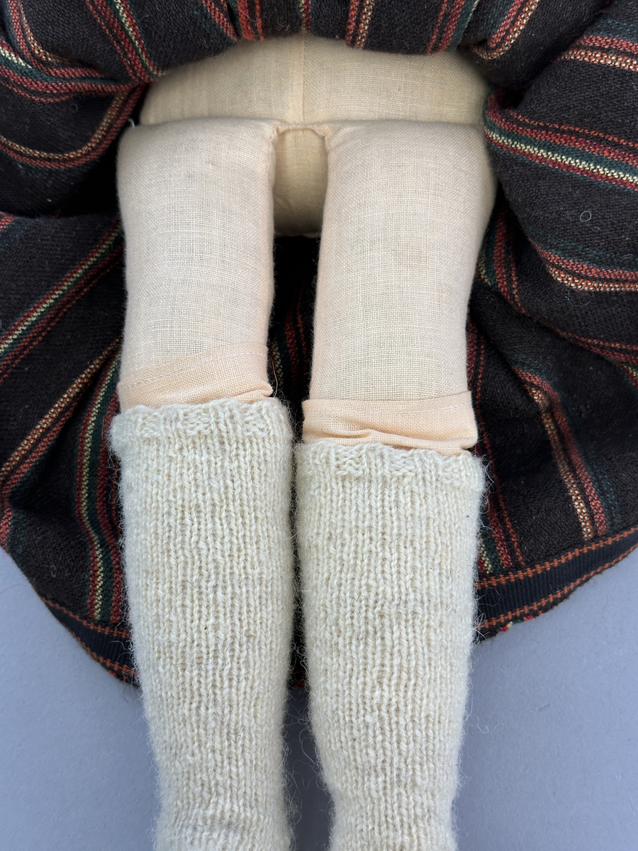 Docka klädd i kvinnlig sockendräkt från Sunne socken i Värmland. Kroppen är handsydd i linnetyg och stoppad med textilt material. Huvud och händer är av lergods. Kläder handsydda av handvävda tyger förutom skjorta, underkjol och halskläde, som är maskinvävt. Skjorta, broderad med stopphålsömsbroderi i vitt lingarn längs sprund mitt fram, har stripade veck runt krage och manschett. Halskläde, broderad med tambursöm i vitt moulinegarn, hålls ihop av en sölja i nysilver med graverat mönster. Yllekjol är rynkad upptill, utom under förkläde, vars linning och skoningsband består av ett bomullsband. Förklädet har lagda veck vid midja fastsydda mot en linning av bomullsband. Livstycket med sköld i ylletyg, är vändsytt/fodrat med ett mönstrat bomullstyg och knäpps med 5 hyskar & hakar samt har 2 små tennknappar fastsydda på utsidan. Ullstrumporna är handstickade i slätstickning med en kant upptill i 2-2 resår. Huvudet är prytt med en vit linnehätta kantad med en knypplad linnespets. Dockans fötter bär svarta skinnskor.