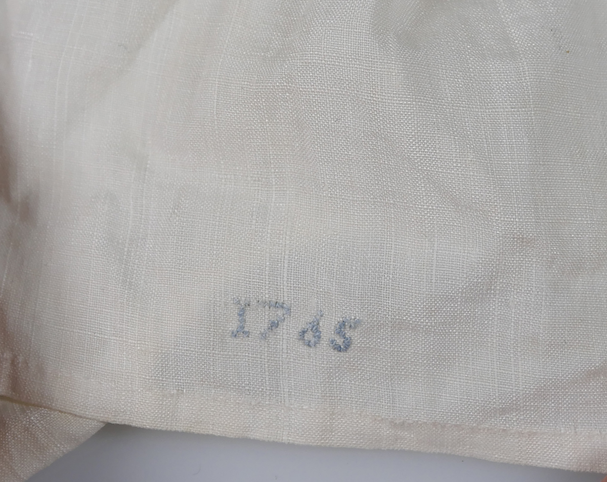 Skjorta i vit bomull, Kantad med spets och med krage och knytning. I nederkant broderat i blått: "AMS N 6 1765"