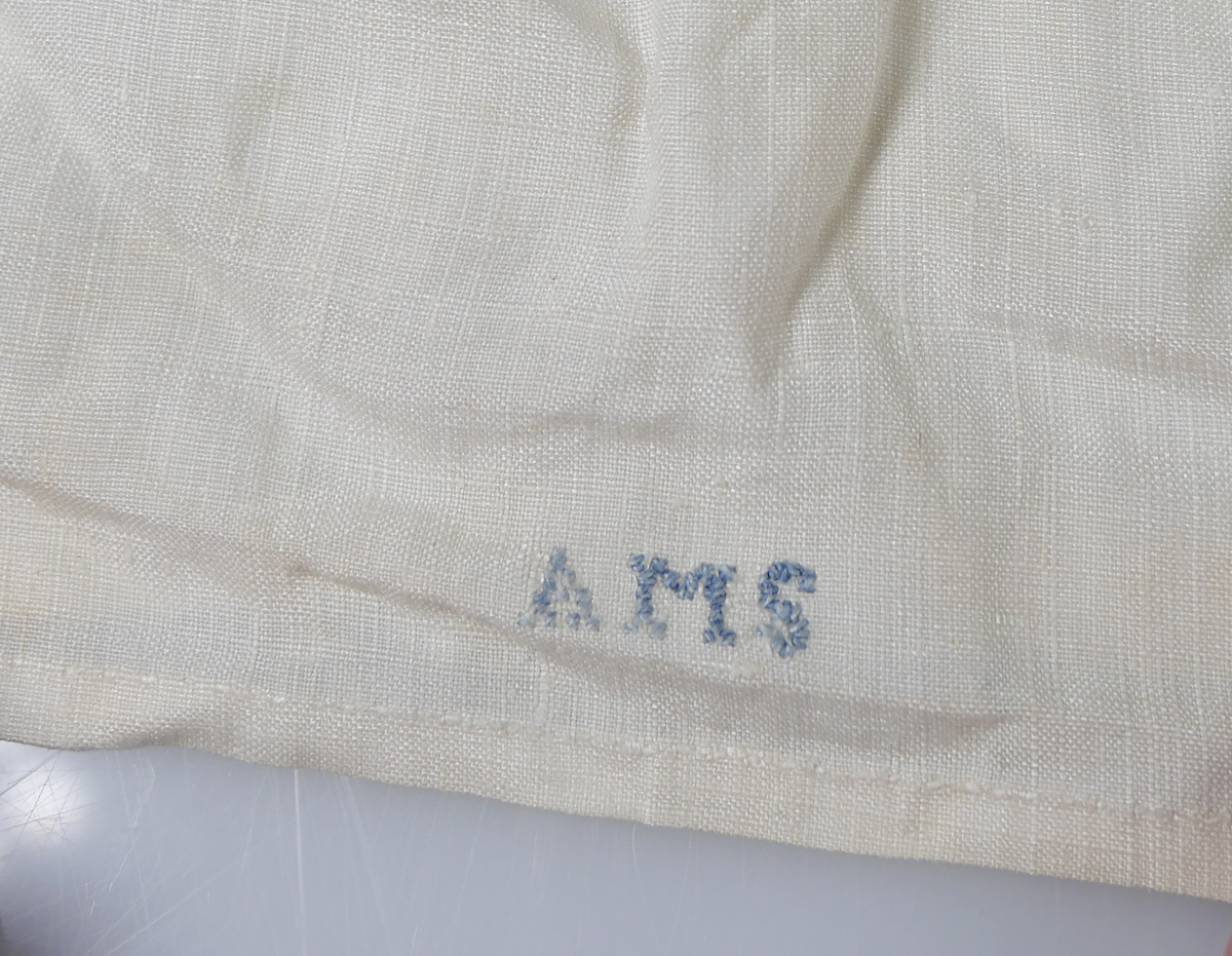 Skjorta i vit bomull, Kantad med spets och med krage och knytning. I nederkant broderat i blått: "AMS N 6 1765"