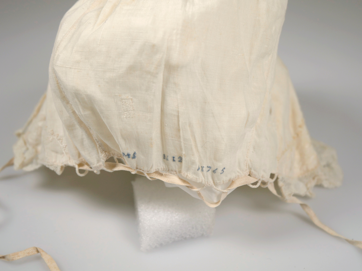 Två spädbarnsmössor av tunn bomull. Båda med spets runt kanten och knytband under hakan. I nacken broderat "AMS N12 A1765" i blått på båda.. 
AM 2024-12 B stora revor i tyget. 