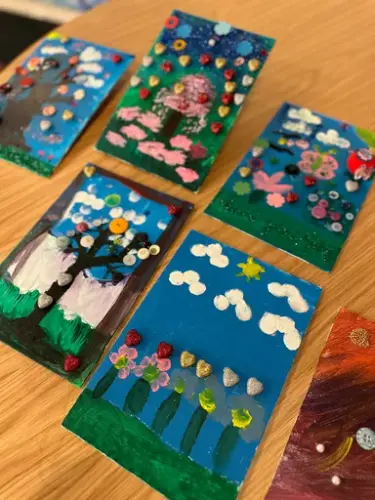 Små fargerike kunstverk laget av barn, med motiver som blomster, trær og skyer, stilt ut på et bord.