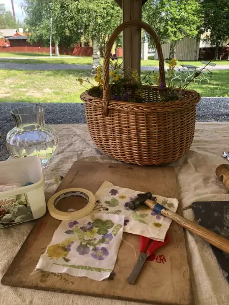Underlägg på ett bord med tyg färgat med blommor, hammare, sax och tejp. Det står en korg med blommor och en vas med vatten på bordet också. I bakgrunden ser man grönt gräs och gröna träd. 