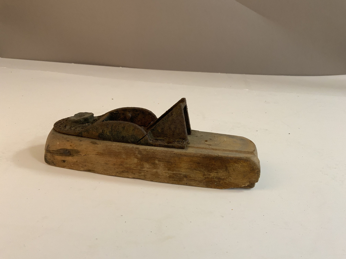 En avrundet treklosse med et hull til eventuelle høvelblad. En jerndel er festet på toppen er brukket og har en som har en hengslet del. Den hengslete delen er brukket og det ser ut som en bit mangler. På ene siden av jerndelen er det spikret fast i treklossen med en lærbit i mellom. Jernbiten ved den hengslete biten var festet med to skruer, hvorav en av de mangler.