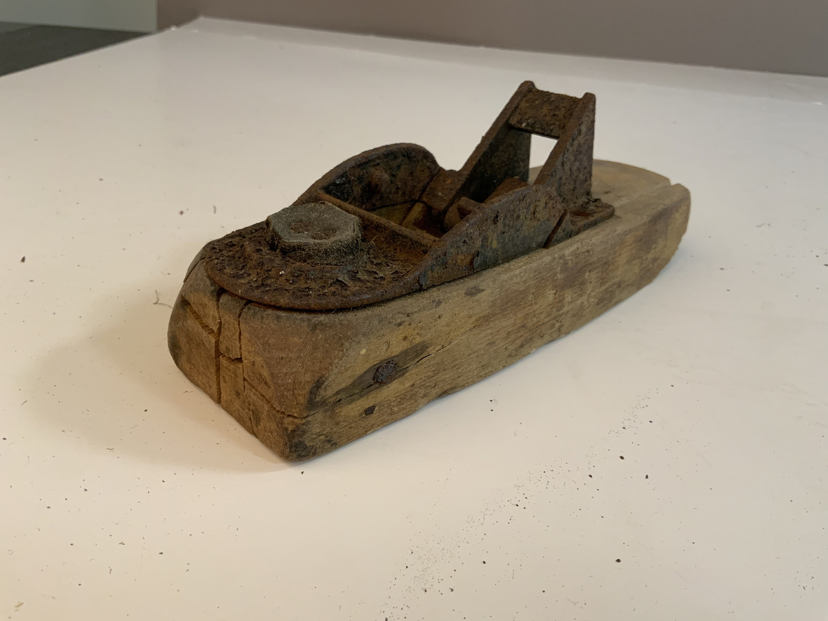 En avrundet treklosse med et hull til eventuelle høvelblad. En jerndel er festet på toppen er brukket og har en som har en hengslet del. Den hengslete delen er brukket og det ser ut som en bit mangler. På ene siden av jerndelen er det spikret fast i treklossen med en lærbit i mellom. Jernbiten ved den hengslete biten var festet med to skruer, hvorav en av de mangler.