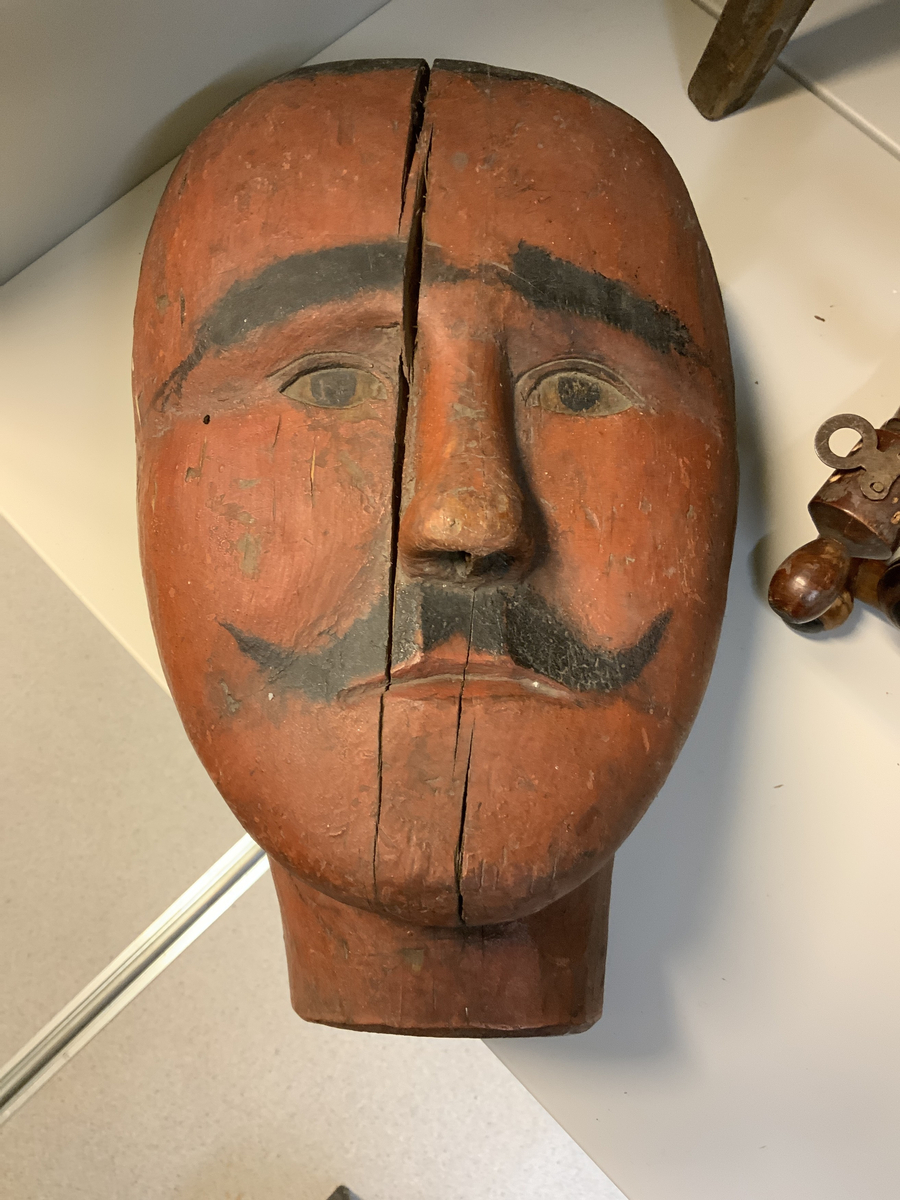 Et mannshode med mustasje. Utskåret, hals, hake, nese og øyne, og påmalt hår, øyebryn, pupiller og mustasje.