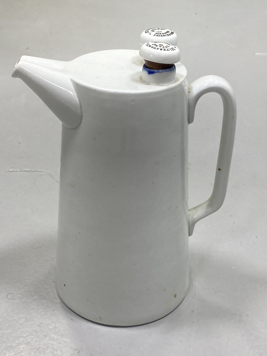 Mugge i glasert keramikk med hanke og helletut. Muggen har to kamre, og helletut med to åpninger, og to påfyllingshull med stoppekorker med gummipakning og hull på toppen.