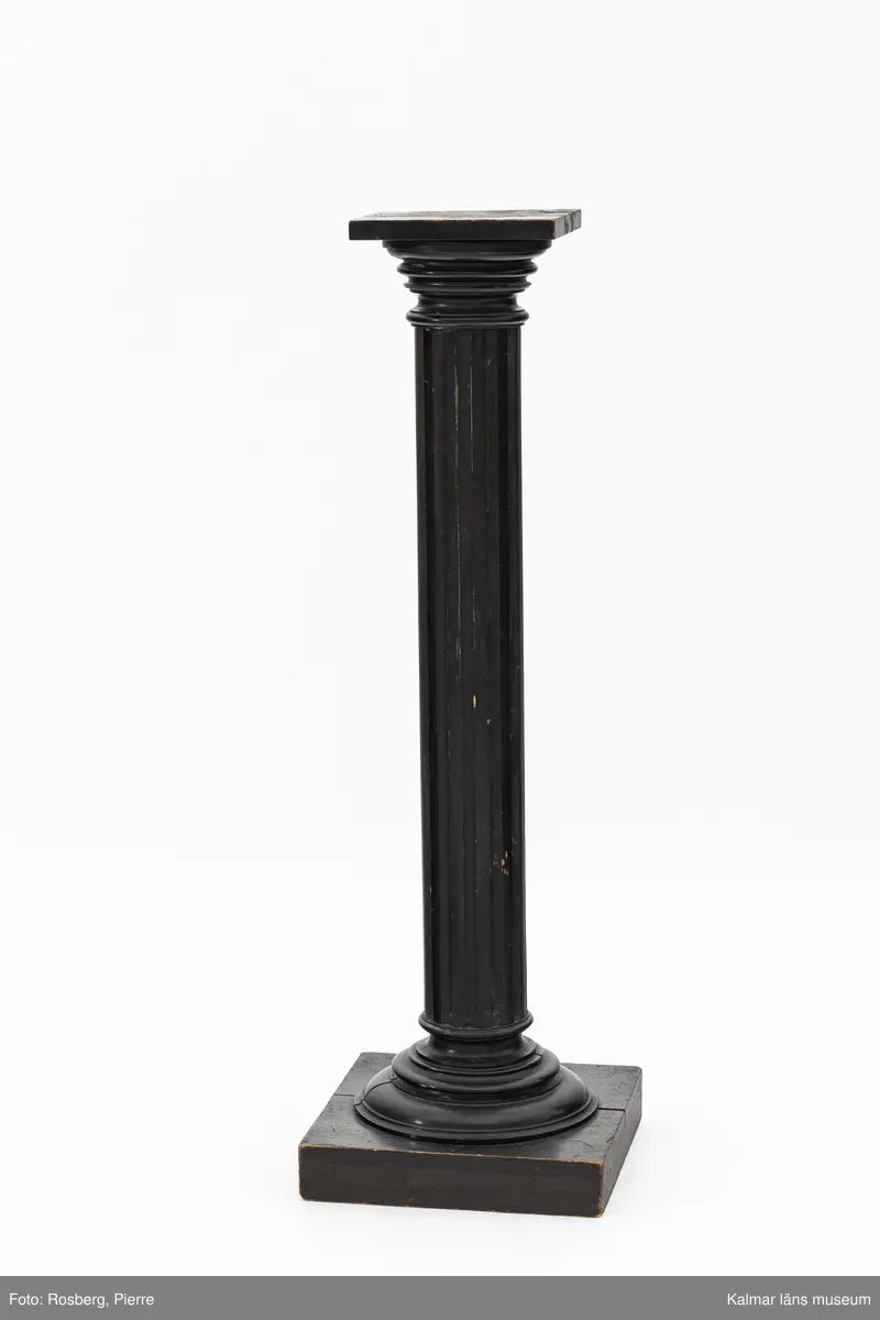 KLM 28467:26. Piedestal. Av trä med svart färg. Formad som en kolonn med kvadratisk överdel och basplatta. Påskrift undersida: Emil Timan Kalmar.