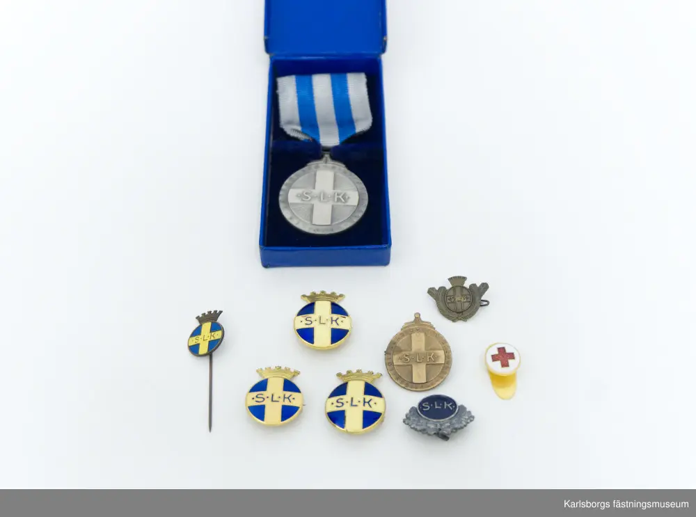 Blå förvaringsask med innehåll av Svenska Lotta Kårens utmärkelser. Tre stycken S.L.K. emblem: runda emblem med en krona överst, gult kors på blå botten och texten "S.L.K.". Säkerhetsnål sitter på baksidan. Två är tillverkade i Stockholm av företaget Sporrong medan den tredje är tillverkad av Westins i Stockholm. S.L.K. Cirkelrund nål av mässing i bronsfärg med "granbarr" runt om, säkerhetsnål på baksidan. Brosch i silver: på dess framsida ses en bädd av eklöv varpå är en cirkel med blå emaljyta. I cirkeln står "S.L.K.". Tre små hål finns längst ner på eklövbäddet, i mellersta hålet hänger en stjärna. Broschens baksida är slät med nålfäste. Tillverkad av Sporrong. Bronsmärke: kors i mitten med texten "S.L.K." och utmed den runda kanten "Riksförbundet Sveriges Lottakårer." Säkerhetsnål på baksidan. Tillverkad av Sporrong. Nålmärke för S.L.K., tillverkad av Sporrong. Knapp i guldfärg, röda korset, tillverkad av Sporrong. Förvaringsasken tillhör förtjänstmedaljen i silver, en lottamedalj. På medaljens framsida i mitten syns ett kors med S.L.K. och kring medaljens kant texten "Riksförbundet Sveriges Lottakårer." På dess baksida sitter ett växtornament runt kanten och i mitten texten "För Förtjänster om Sveriges Lottaorganisation" samt stämpeltecken. Medaljen är fäst i ett band: vitt med två blå ränder, på baksidan säkerhetsnål för fastsättning.