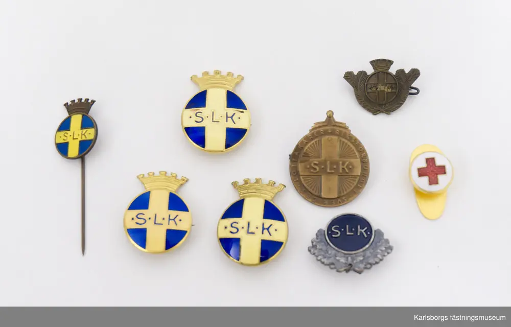 Blå förvaringsask med innehåll av Svenska Lotta Kårens utmärkelser. Tre stycken S.L.K. emblem: runda emblem med en krona överst, gult kors på blå botten och texten "S.L.K.". Säkerhetsnål sitter på baksidan. Två är tillverkade i Stockholm av företaget Sporrong medan den tredje är tillverkad av Westins i Stockholm. S.L.K. Cirkelrund nål av mässing i bronsfärg med "granbarr" runt om, säkerhetsnål på baksidan. Brosch i silver: på dess framsida ses en bädd av eklöv varpå är en cirkel med blå emaljyta. I cirkeln står "S.L.K.". Tre små hål finns längst ner på eklövbäddet, i mellersta hålet hänger en stjärna. Broschens baksida är slät med nålfäste. Tillverkad av Sporrong. Bronsmärke: kors i mitten med texten "S.L.K." och utmed den runda kanten "Riksförbundet Sveriges Lottakårer." Säkerhetsnål på baksidan. Tillverkad av Sporrong. Nålmärke för S.L.K., tillverkad av Sporrong. Knapp i guldfärg, röda korset, tillverkad av Sporrong. Förvaringsasken tillhör förtjänstmedaljen i silver, en lottamedalj. På medaljens framsida i mitten syns ett kors med S.L.K. och kring medaljens kant texten "Riksförbundet Sveriges Lottakårer." På dess baksida sitter ett växtornament runt kanten och i mitten texten "För Förtjänster om Sveriges Lottaorganisation" samt stämpeltecken. Medaljen är fäst i ett band: vitt med två blå ränder, på baksidan säkerhetsnål för fastsättning.