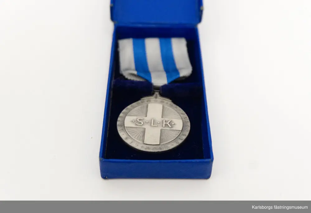 Blå förvaringsask med innehåll av Svenska Lotta Kårens utmärkelser. Tre stycken S.L.K. emblem: runda emblem med en krona överst, gult kors på blå botten och texten "S.L.K.". Säkerhetsnål sitter på baksidan. Två är tillverkade i Stockholm av företaget Sporrong medan den tredje är tillverkad av Westins i Stockholm. S.L.K. Cirkelrund nål av mässing i bronsfärg med "granbarr" runt om, säkerhetsnål på baksidan. Brosch i silver: på dess framsida ses en bädd av eklöv varpå är en cirkel med blå emaljyta. I cirkeln står "S.L.K.". Tre små hål finns längst ner på eklövbäddet, i mellersta hålet hänger en stjärna. Broschens baksida är slät med nålfäste. Tillverkad av Sporrong. Bronsmärke: kors i mitten med texten "S.L.K." och utmed den runda kanten "Riksförbundet Sveriges Lottakårer." Säkerhetsnål på baksidan. Tillverkad av Sporrong. Nålmärke för S.L.K., tillverkad av Sporrong. Knapp i guldfärg, röda korset, tillverkad av Sporrong. Förvaringsasken tillhör förtjänstmedaljen i silver, en lottamedalj. På medaljens framsida i mitten syns ett kors med S.L.K. och kring medaljens kant texten "Riksförbundet Sveriges Lottakårer." På dess baksida sitter ett växtornament runt kanten och i mitten texten "För Förtjänster om Sveriges Lottaorganisation" samt stämpeltecken. Medaljen är fäst i ett band: vitt med två blå ränder, på baksidan säkerhetsnål för fastsättning.