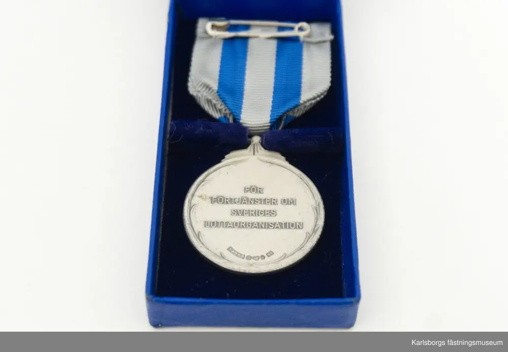 Blå förvaringsask med innehåll av Svenska Lotta Kårens utmärkelser. Tre stycken S.L.K. emblem: runda emblem med en krona överst, gult kors på blå botten och texten "S.L.K.". Säkerhetsnål sitter på baksidan. Två är tillverkade i Stockholm av företaget Sporrong medan den tredje är tillverkad av Westins i Stockholm. S.L.K. Cirkelrund nål av mässing i bronsfärg med "granbarr" runt om, säkerhetsnål på baksidan. Brosch i silver: på dess framsida ses en bädd av eklöv varpå är en cirkel med blå emaljyta. I cirkeln står "S.L.K.". Tre små hål finns längst ner på eklövbäddet, i mellersta hålet hänger en stjärna. Broschens baksida är slät med nålfäste. Tillverkad av Sporrong. Bronsmärke: kors i mitten med texten "S.L.K." och utmed den runda kanten "Riksförbundet Sveriges Lottakårer." Säkerhetsnål på baksidan. Tillverkad av Sporrong. Nålmärke för S.L.K., tillverkad av Sporrong. Knapp i guldfärg, röda korset, tillverkad av Sporrong. Förvaringsasken tillhör förtjänstmedaljen i silver, en lottamedalj. På medaljens framsida i mitten syns ett kors med S.L.K. och kring medaljens kant texten "Riksförbundet Sveriges Lottakårer." På dess baksida sitter ett växtornament runt kanten och i mitten texten "För Förtjänster om Sveriges Lottaorganisation" samt stämpeltecken. Medaljen är fäst i ett band: vitt med två blå ränder, på baksidan säkerhetsnål för fastsättning.