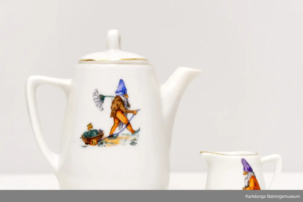 Dockservis av porslin bestående av kaffekanna, mjölkkanna och en tallrik för kannan. Kaffekanna: 11,5cm hög. Mjölkkanna: ca 3,3cm hög. Tallrikens diameter: 6,5cm. Porslinets motiv skildrar sagoscener: små tomtefigurer där en drar en vagn med liten sittandes pojke med krona. Den andra tomtefiguren sitter på en svamp och framför sig en kråka, eller korp, bärandes på en krona och bär med näbben en ring. Det finns även en figur liknande en älva, i vagn av blad och blomma som dras av en fågel. Hon är naken med vita vingar och krona där ett kors sitter längst upp. Guldmönstrad kantbård finns på alla tre porslin-ting.