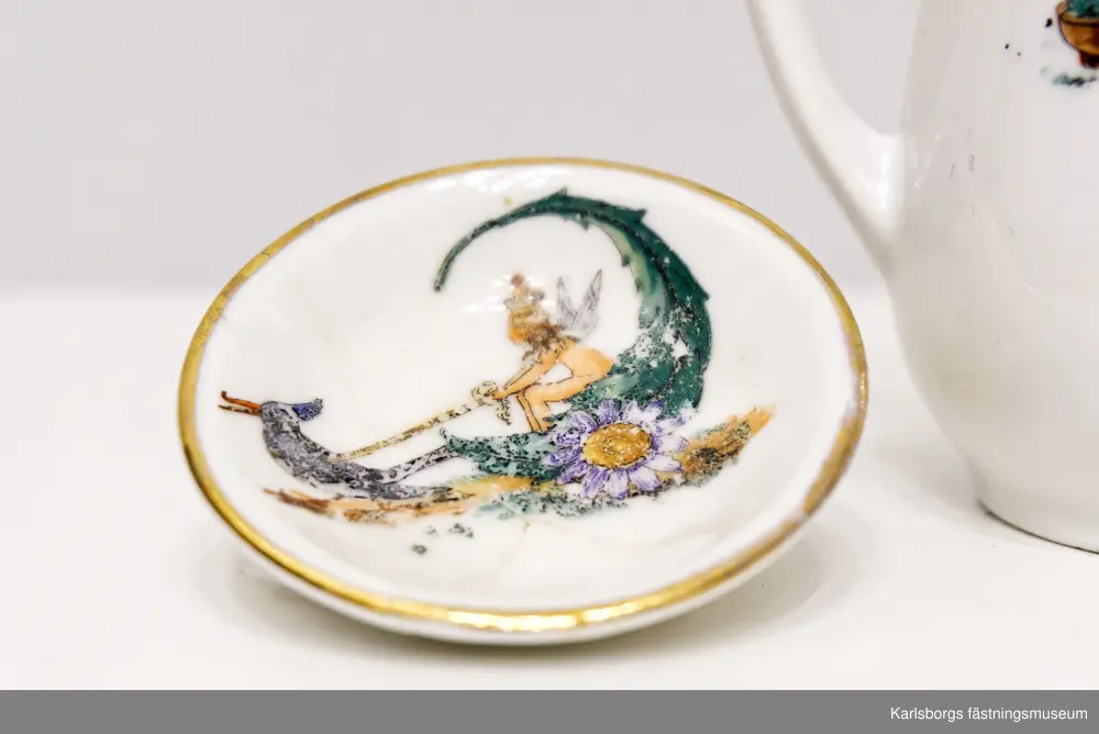 Dockservis av porslin bestående av kaffekanna, mjölkkanna och en tallrik för kannan. Kaffekanna: 11,5cm hög. Mjölkkanna: ca 3,3cm hög. Tallrikens diameter: 6,5cm. Porslinets motiv skildrar sagoscener: små tomtefigurer där en drar en vagn med liten sittandes pojke med krona. Den andra tomtefiguren sitter på en svamp och framför sig en kråka, eller korp, bärandes på en krona och bär med näbben en ring. Det finns även en figur liknande en älva, i vagn av blad och blomma som dras av en fågel. Hon är naken med vita vingar och krona där ett kors sitter längst upp. Guldmönstrad kantbård finns på alla tre porslin-ting.