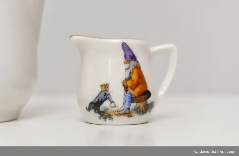 Dockservis av porslin bestående av kaffekanna, mjölkkanna och en tallrik för kannan. Kaffekanna: 11,5cm hög. Mjölkkanna: ca 3,3cm hög. Tallrikens diameter: 6,5cm. Porslinets motiv skildrar sagoscener: små tomtefigurer där en drar en vagn med liten sittandes pojke med krona. Den andra tomtefiguren sitter på en svamp och framför sig en kråka, eller korp, bärandes på en krona och bär med näbben en ring. Det finns även en figur liknande en älva, i vagn av blad och blomma som dras av en fågel. Hon är naken med vita vingar och krona där ett kors sitter längst upp. Guldmönstrad kantbård finns på alla tre porslin-ting.