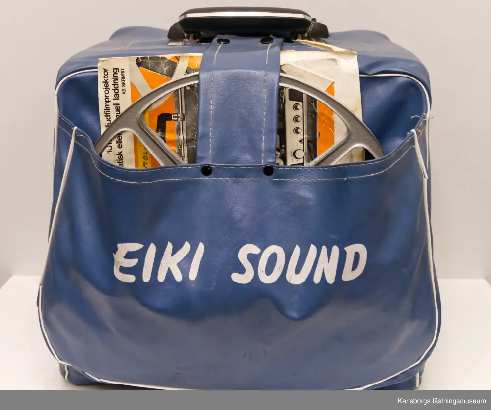 Eiki serie ST 39573, 16 mm ljudfilmprojektor med både automatisk och manuell laddning. Till projektorn hör även en anslutningskabel, filmrulle och lampa. Projektorn skyddas av ett blått överdrag.