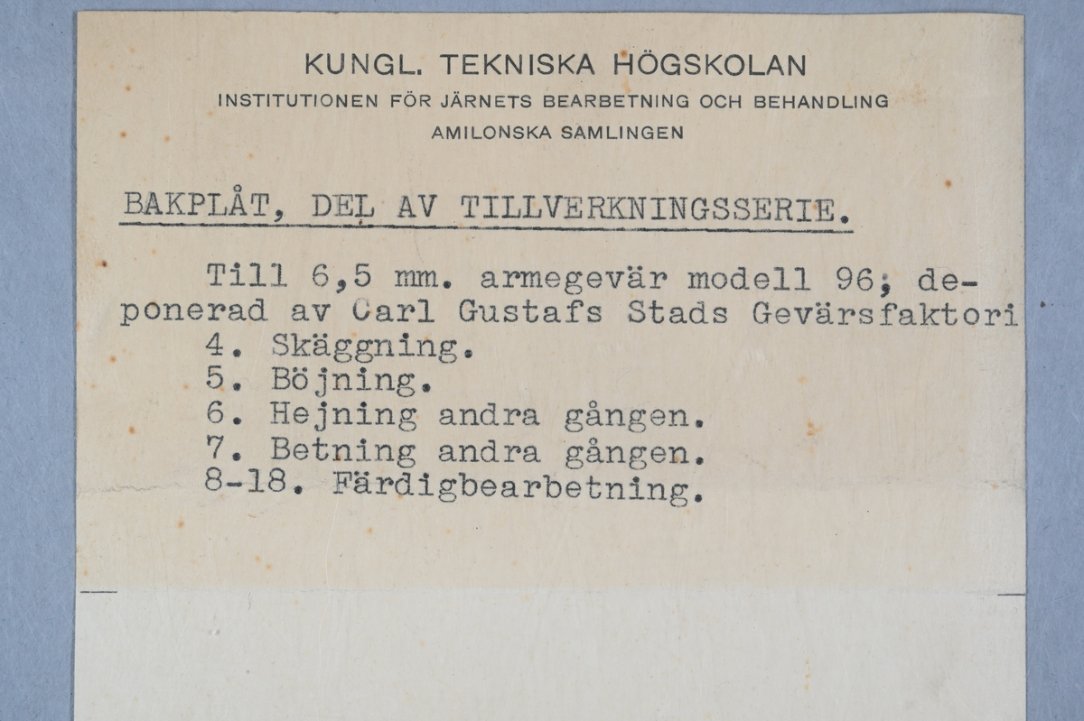Fem prover på bakplåt till armegevär. Märkning på proverna med tilldelade siffror: 4, 5, 6, 7 och 18. Tillhörande lapp med notering: "Bakplåt, del av tillverkningsserie. Till 6,5 mm. armegevär modell 96, deponerad av Carl Gustafs Stads Gervärsfaktori. 
4. Skäggning.
5. Böjning.
6. Hejning andra gången.
7. Betning andra gången.
8-18. Färdigbearbetning."