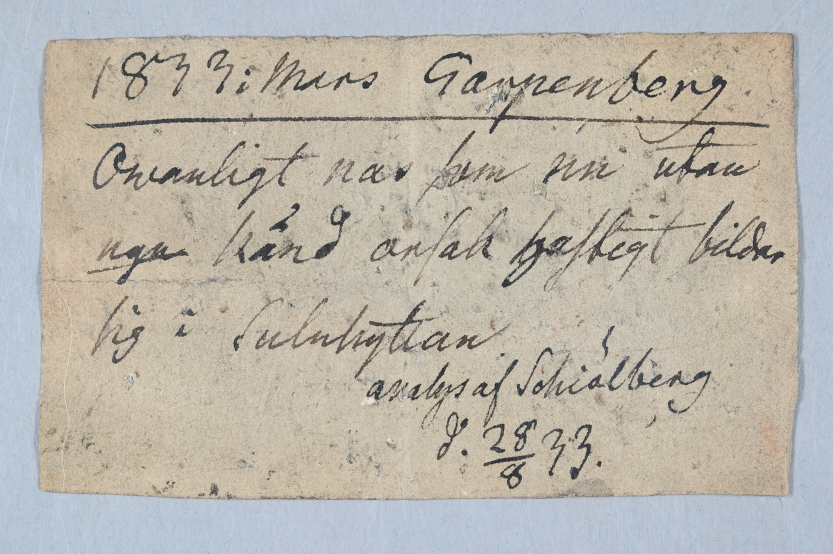 Materialprov, Grå, blå och brunaktig. Tillhörande lapp med notering: "1833 i Mars Garpenberg. Owanligt nas som nu utan ugn bränd ansatt hastigt bildar sig i suluhyttan. Analys as Schiälberg (?) d. 28/8 33."