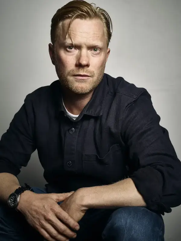 Portrettfoto av Leander Djønne med mørk skjorte sitjande framom ein grå vegg